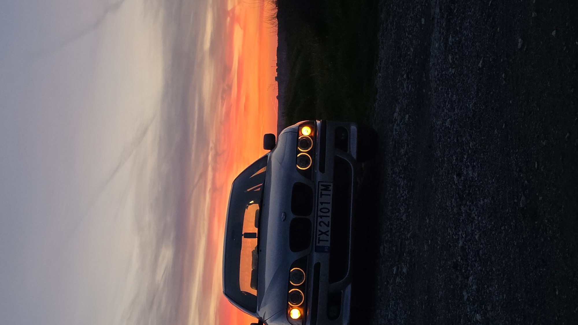 The image captures the front view of a silver or light grey BMW 5 Series (E39 model), parked on a dark, gravelly road or surface. The car is angled slightly, with its front right side facing closer to the viewer. Its distinctive "angel eyes" headlights are illuminated, casting a warm, circular glow.
Behind the car, a dramatic sunset dominates the background, filling the sky with vibrant and intense colors. Streaks of fiery orange, deep red, and purple hues transition into lighter shades towards the upper left, indicating twilight or dusk. A dark, silhouetted horizon line, possibly composed of distant hills or land with sparse trees, separates the car from the colorful sky.
The scene depicts a peaceful moment during sunset in what appears to be a rural or open landscape, consistent with the given location context of Balgarevo, Bulgaria. The vibrant sky provides a striking contrast to the darker foreground and the car itself. No individuals are visible in the frame.
A notable detail is the car's front license plate, which displays the registration "TX 2101 TM" and includes a small "BG" emblem, confirming its Bulgarian origin. The time of day is clearly sunset, creating a captivating and atmospheric visual.