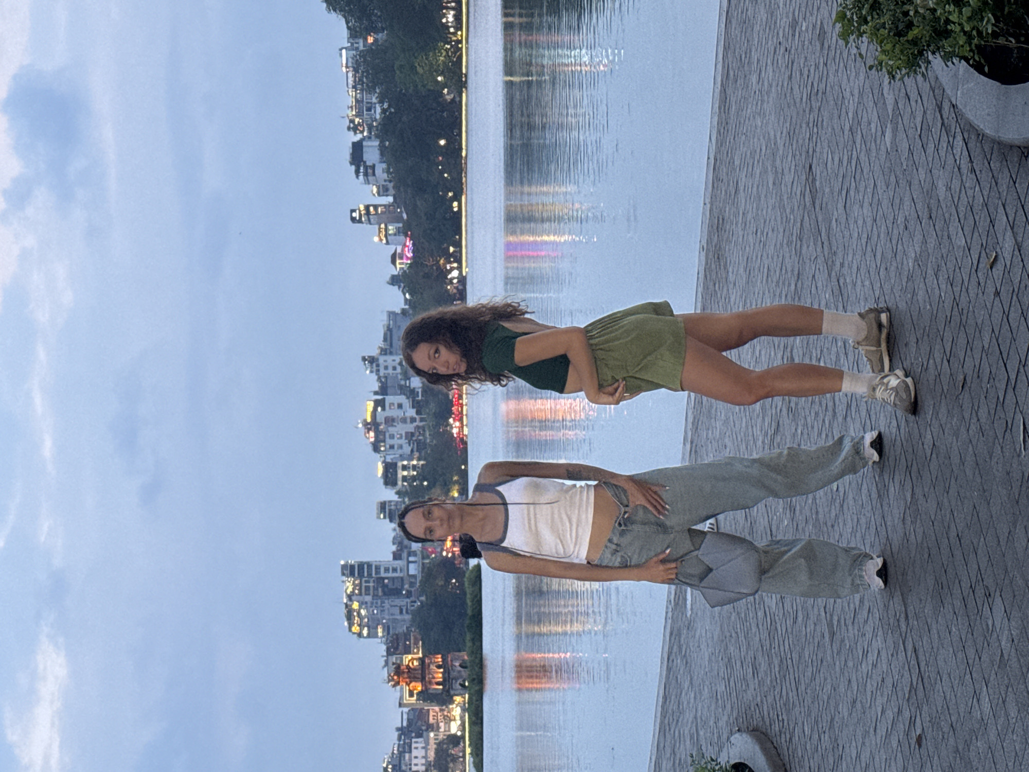 Two young women stand on a paved walkway beside a body of water, likely Hoàn Kiếm Lake in Hanoi, Vietnam. The woman closer to the camera wears a white tank top and light blue, baggy jeans, and a shoulder bag. The other woman wears a green two-piece outfit. Buildings are seen across the water. The time is likely evening due to the sky's lighting and reflections of city lights on the water.