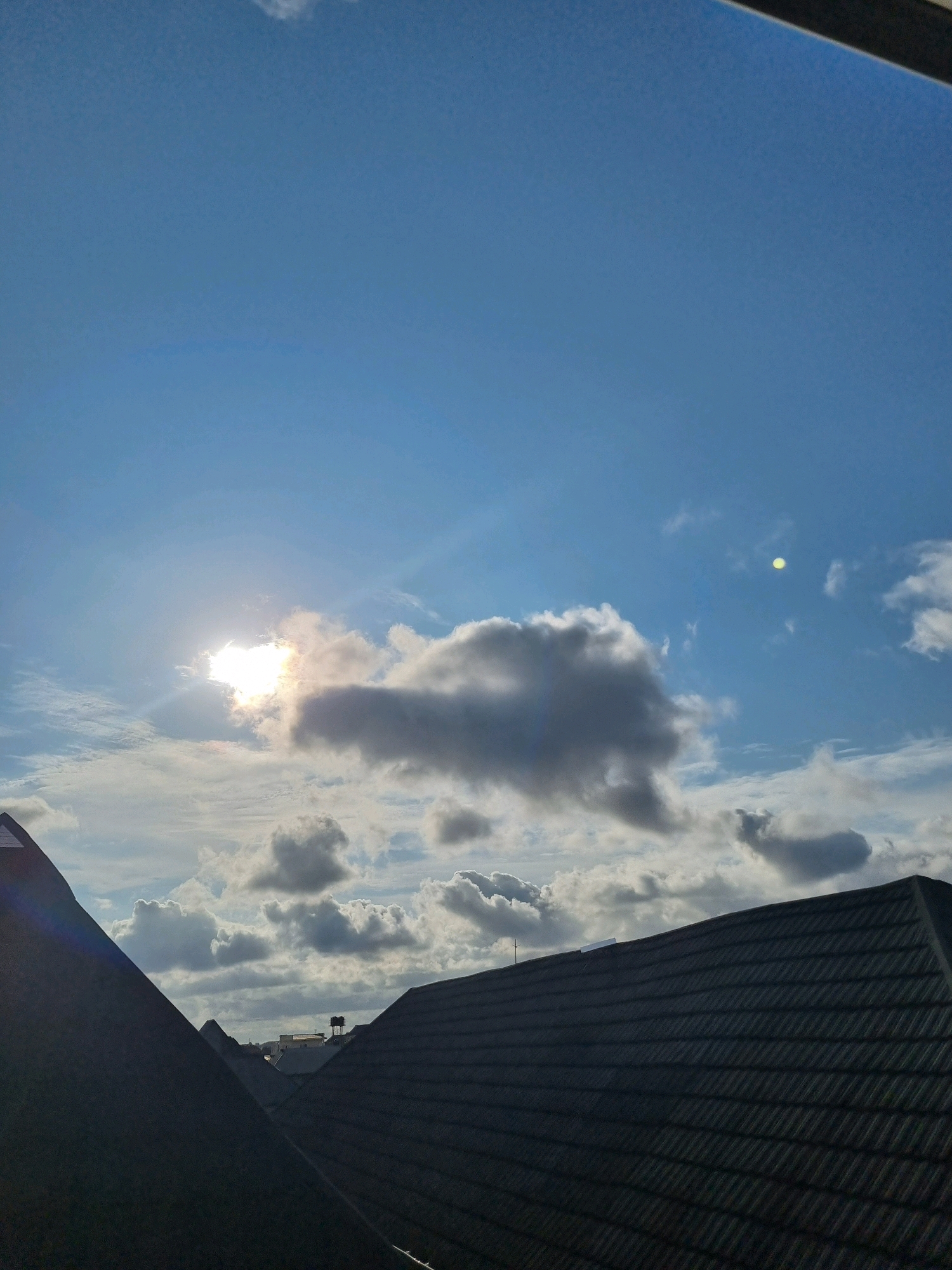 The image shows a view of rooftops against a bright blue sky with scattered clouds. The sun is partially obscured by a large, dark cloud in the center left of the frame, creating a lens flare effect. Another small, bright circular object, possibly another sun or a distant light source, is visible in the upper right quadrant of the sky. The foreground is dominated by the dark, angled rooftops of buildings, suggesting an urban or suburban environment. The overall impression is of a sunny day with some cloud cover, possibly late morning or early afternoon given the sun's position.