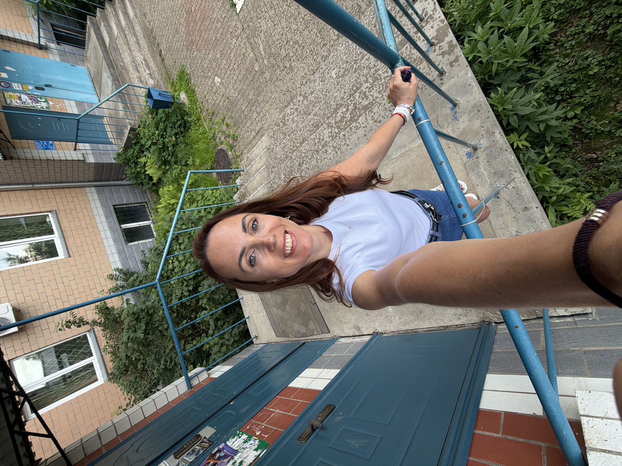 A woman with long, brown hair and blue eyes smiles as she takes a selfie on a set of outdoor stairs. She is wearing a white t-shirt, blue denim shorts, and white sneakers. She has a watch and a bracelet on her left wrist. The stairs are made of concrete and have a blue metal railing. The building behind her is made of brick and has blue doors. There is green foliage growing around the base of the stairs and along the side of the building. The image appears to have been taken on a cloudy day. The location is Birobidzhan, Russia.