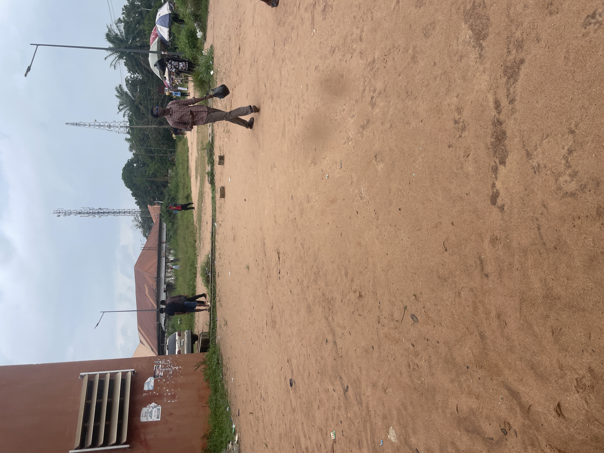 An outdoor, daytime scene depicts a dirt lot in Ihiagwa, Nigeria. In the foreground, a man walks towards the right, carrying a black plastic bag and wearing a plaid shirt and dark pants. A second man, dressed in a black t-shirt and dark shorts, stands to the left in the middle ground, next to another man wearing a dark shirt and patterned shorts. A gold-colored SUV is parked behind them. In the background, a building with a reddish-orange facade and a louvered window is visible on the far left. Beyond this, a series of buildings with red roofs are present, along with tall communication towers and lush green trees. Several market stalls with umbrellas are set up further in the background on the right. The ground is covered in reddish-brown dirt with scattered debris. The sky is overcast.