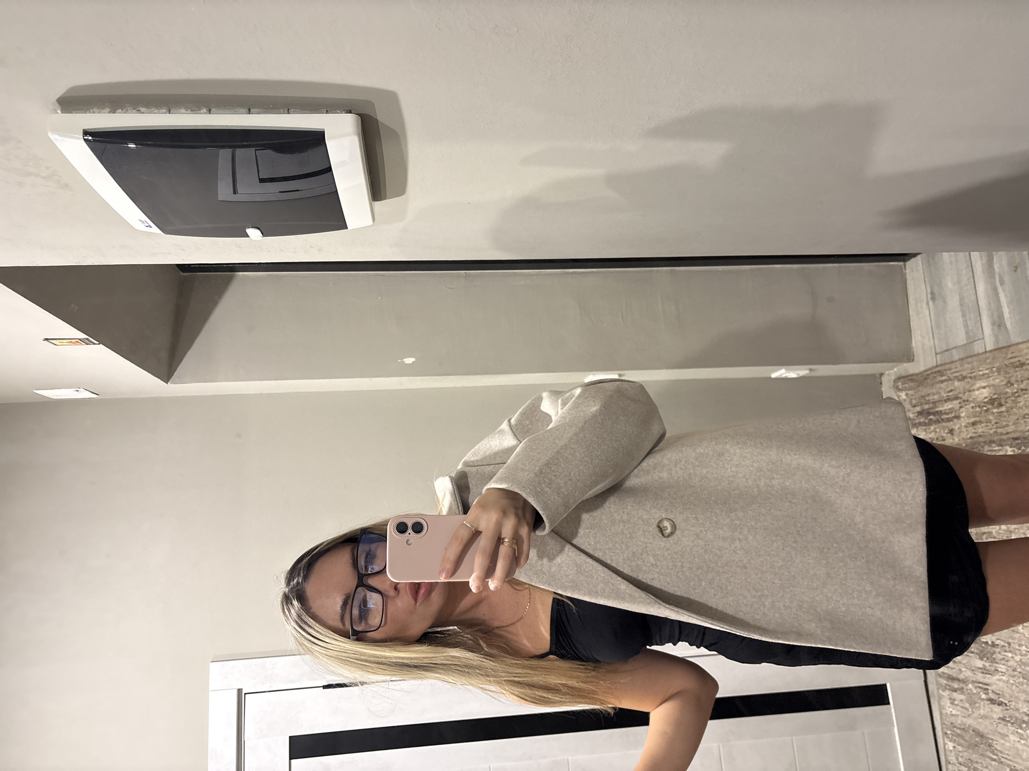 The image captures a female subject taking a mirror selfie indoors, located in the city of Votkinsk, Russia. The subject, possessing long blonde hair and wearing black-rimmed glasses, is dressed in a black strapless top or dress and a thin gold-colored necklace. A light beige double-breasted coat is draped over her left shoulder and arm, with two visible buttons on the coat. She has at least two gold-colored rings on her left hand. The subject is holding a pink smartphone in her right hand, angled to capture her reflection. The background features a white door with a prominent black vertical stripe on the left side. To the right, a light grey wall displays a wall-mounted white and black dispenser or panel, possibly an elevator call unit or a sanitary dispenser, with a small white lever/button. A partial view of a small red rectangular sign is visible above this panel. The floor consists of light grey tiles and a textured, light brown/beige rug or mat.