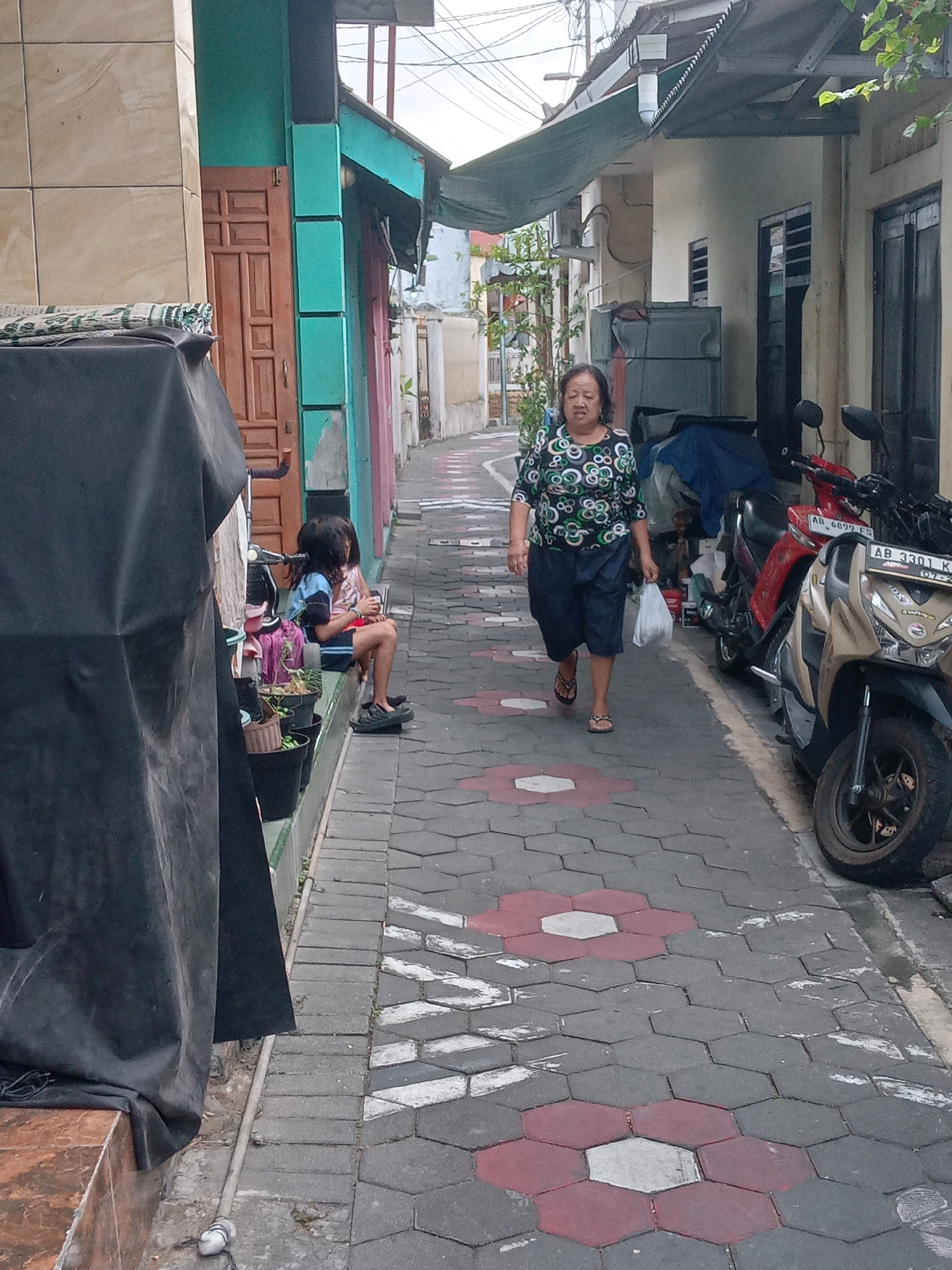 An elderly Indonesian woman with dark hair is walking down a narrow, paved alleyway in Yogyakarta, Indonesia. She is wearing a colorful, circular-patterned shirt and dark blue shorts, and she is carrying a white plastic bag. The alleyway is lined with buildings on both sides. On the left, there is a building with a light brown tiled facade and a turquoise section with a wooden door. A black tarp is draped over something in the foreground on the left. On the right side of the alley, there are houses with light-colored walls and dark windows. Several motorcycles are parked along the right side of the alley. Two children are sitting on a raised concrete ledge on the left side of the alley, next to potted plants. The pavement in the alleyway is made of hexagonal interlocking stones, some of which are decorated with red and white flower-like patterns. The sky is overcast.
