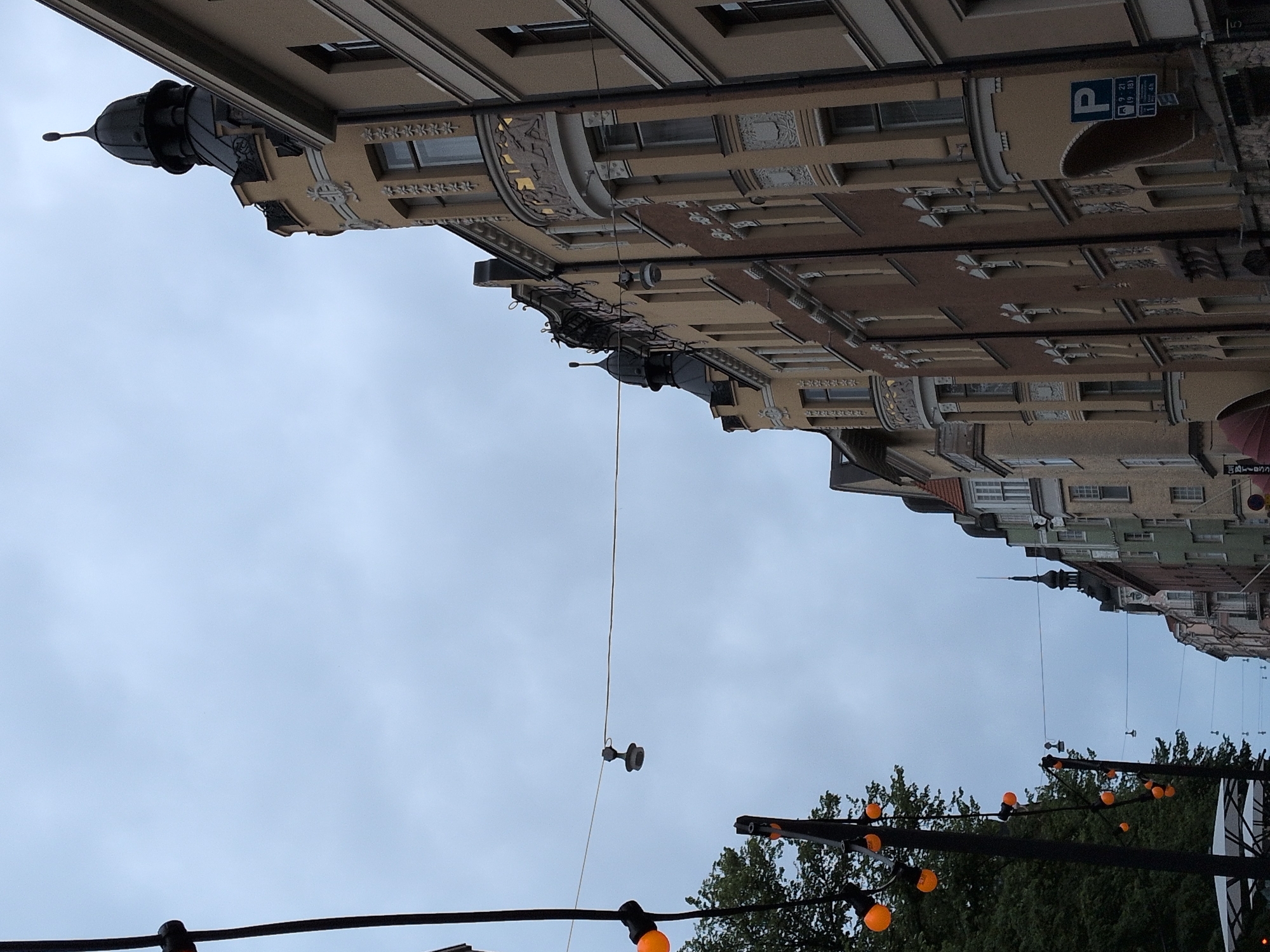 The image shows a view of a street in Helsinki, Finland, under a cloudy sky. On the right side, several multi-story buildings with ornate facades line the street. These buildings feature different architectural styles, with some having detailed carvings above the windows and rounded balconies. One building on the far right has a parking sign with Finnish text and symbols, indicating parking regulations. On the left side of the image, a tree-lined area is visible, and strings of orange decorative lights are suspended across the street between poles. Some of these lights are illuminated. A thin wire stretches across the sky, with a single, dark-colored lamp hanging from it. The overall scene suggests an urban environment, possibly a pedestrianized street or a street with limited vehicle traffic, decorated for an event or with permanent lighting features.