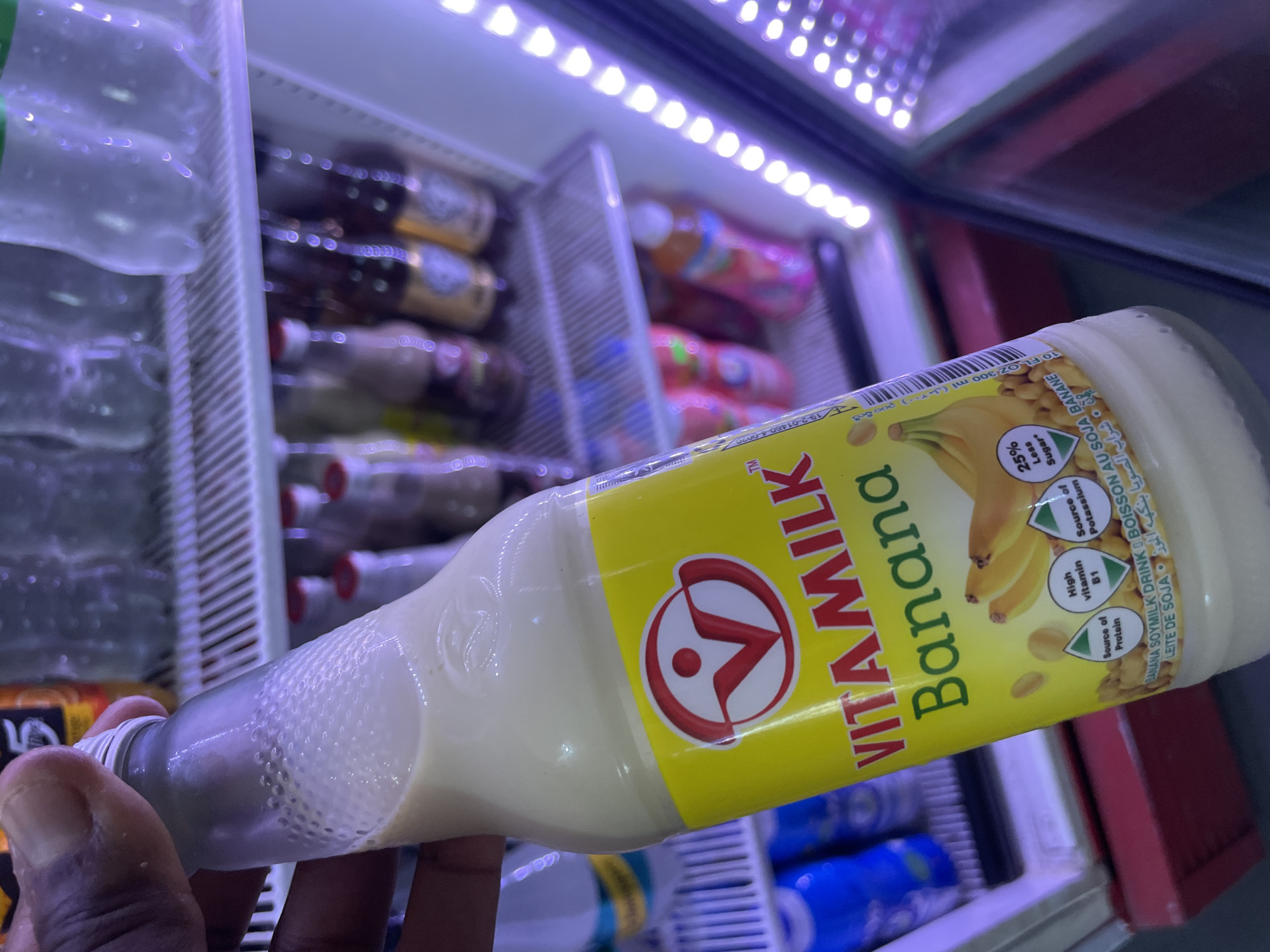 A bottle of Vitamilk Banana Soymilk Drink is being held in front of a refrigerated display case. The bottle's label is predominantly yellow with the words "VITA MILK" in large, bold, red letters. "Banana" is printed in a large, sans-serif font alongside an image of bananas. The label also includes nutritional information, such as being a source of protein, potassium, vitamin B1, and containing 25% less sugar. The bottle is likely made of glass due to the textured appearance near the top. A dark-skinned hand is holding the bottle. The refrigerated display case behind the bottle contains various drinks. The top of the case has LED lighting strips. The image was captured under fluorescent lighting. Given that the brand, Vitamilk, is widely available in Nigeria, the photo could have been taken in Kurunduma, Nigeria, as specified. However, without further verifiable information, the location cannot be definitively confirmed.