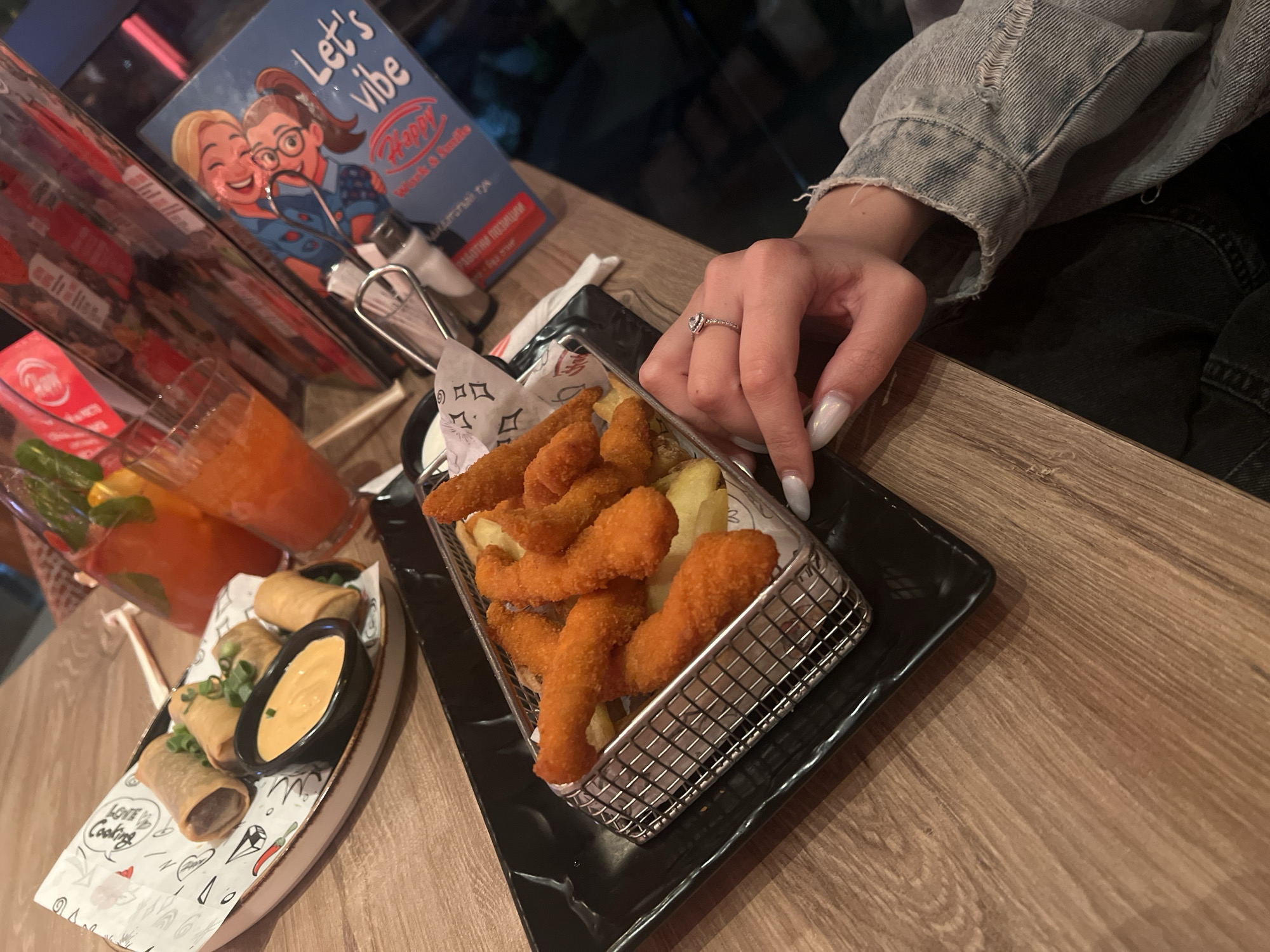 The image captures a close-up view of a dining table scene in what appears to be a restaurant. A person wearing a denim jacket is seated at the table. The person's hand, adorned with a ring, rests near a serving of fried food, possibly chicken strips or similar, presented in a metal basket with fries on a black rectangular plate. To the left of the basket is a plate of spring rolls with a dipping sauce, resting on a decorative paper liner. In the background, there is a beverage, likely a cocktail or juice, in a glass, along with menus or advertising material for "Happy Bar & Grill," a chain restaurant known to exist in Sofia, Bulgaria, among other locations. The table has a wood-grain surface. The lighting suggests an indoor setting. The image focuses on the food and the hand, implying a dining experience. No explicit action other than presence at the table is shown. The environment is a typical restaurant setting, presumably at Happy Bar & Grill in Sofia, Bulgaria, based on the promotional material.