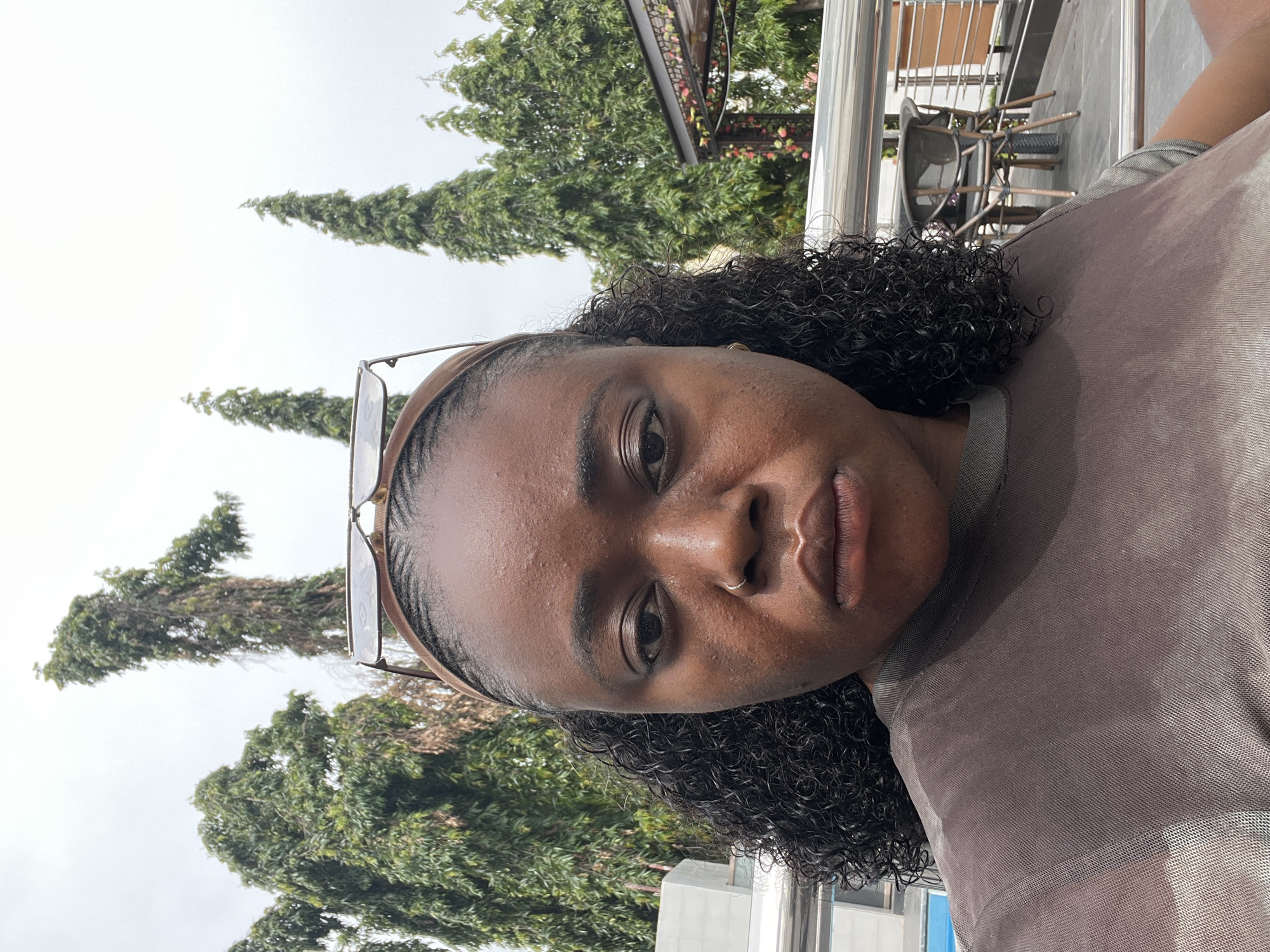 A close-up, medium shot captured in Abuja, Nigeria, shows a dark-skinned woman with curly black hair, wearing a brown headband with sunglasses perched on top. She has a nose ring and is looking directly at the camera with a serious expression. Her skin appears slightly shiny, possibly from sweat. She is wearing a brown, sheer-brushed t-shirt. In the background, lush green trees tower over a building with an outdoor seating area featuring tables and chairs. The sky is overcast, suggesting a cloudy day. The woman is positioned centrally in the frame, with the natural elements of the environment framing her face.
