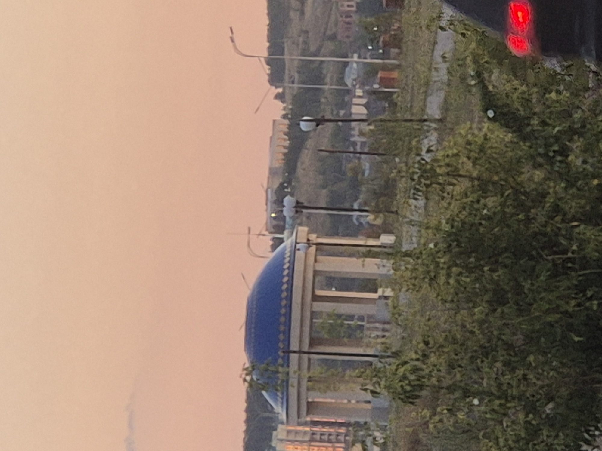 The image depicts an outdoor scene with a prominent gazebo featuring a blue, domed roof adorned with gold accents. The gazebo has a classical design with multiple columns supporting the roof. Surrounding the gazebo are green bushes and trees, indicating a park or natural area. In the background, a city landscape is visible, including buildings and a hillside. The sky is a soft, peachy hue, suggesting it is either sunrise or sunset. The overall atmosphere is serene. The location is identified as the Park of Lovers in Kokshetau, Kazakhstan.