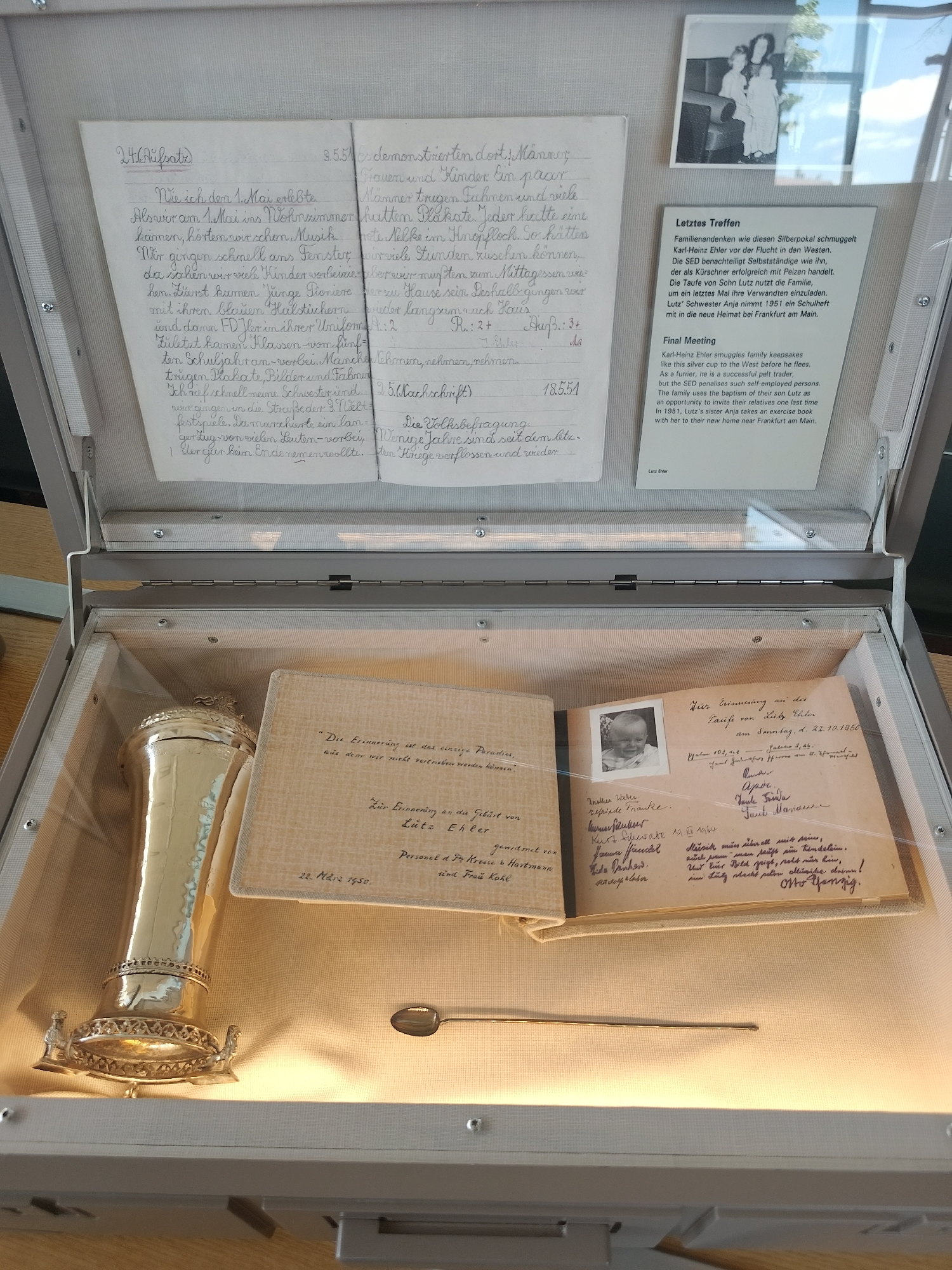 A display case contains various items, including handwritten documents, a silver chalice, a spoon, and photographs. The handwritten documents in German appear to be essays, personal notes, and commemorative texts related to a person named Lutz Ehler and family gatherings. The silver chalice is ornate. A spoon lies next to the notebook. Photographs depict individuals, possibly family members, and events such as a baptism. The contents suggest personal memories and historical documentation, possibly related to East Germany.