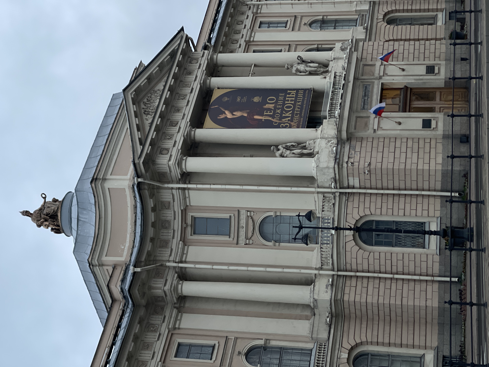 This is a frontal, slightly low-angle exterior shot of the Imperial Academy of Arts in Saint Petersburg, Russia. The building is grand and neoclassical, with a light pink facade and large, cream-colored Corinthian columns. The sky is overcast and a muted grey.
Dominating the center of the building's facade is a large, dark banner with Cyrillic text and an image of a muscular, nude male figure in a dynamic pose. The banner appears to advertise an exhibition.
Above the entrance, flanking the banner, are two classical sculptures of male figures. On the very top of the building, on a rounded dome, sits a large bronze sculpture of a seated female figure holding a wreath, with smaller figures at her feet.
The entrance itself features large, double wooden doors flanked by two smaller Russian flags on flagpoles. Above the entrance, there is architectural detailing and two plaques.
On the right side of the image, towards the bottom, a black lamppost with two lights is visible. A low, black metal fence runs along the bottom of the image, separating the sidewalk from the building. The ground in front of the building appears to be a sidewalk and a street, with a dark road surface.
The overall impression is one of historical grandeur and cultural significance, typical of prominent institutions in Saint Petersburg. The lighting is soft and diffused due to the cloudy weather.