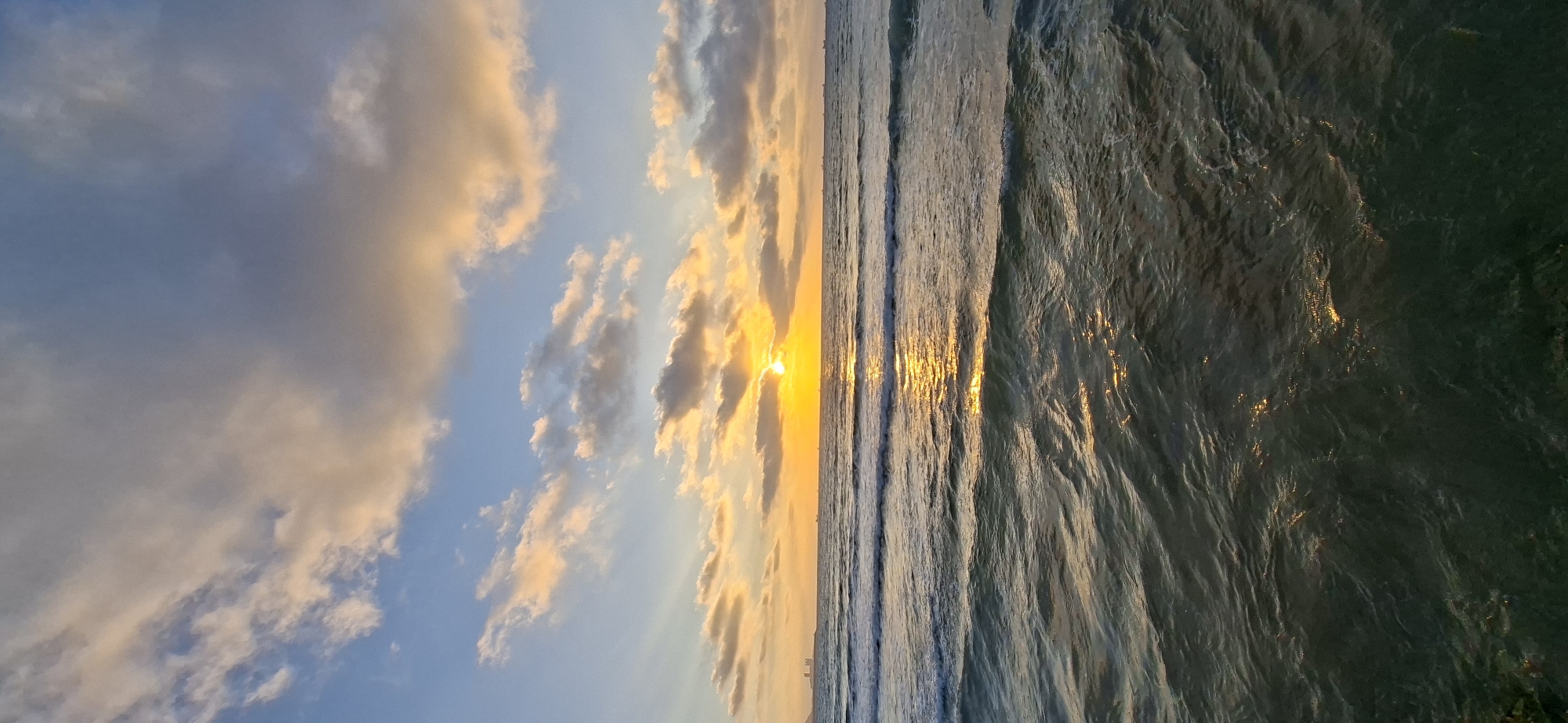 This image depicts a sunset over a body of water. The sun is low on the horizon, casting a warm, golden light across the scene. The sky is a mix of blue and orange hues, with scattered clouds tinged with pink and gold. Gentle waves are rolling in from the sea, reflecting the sunset's glow on their surfaces. The water transitions from a lighter, sunlit area near the horizon to a darker, deeper color in the foreground. The overall mood of the image is serene and peaceful. There is no visible text in the image.