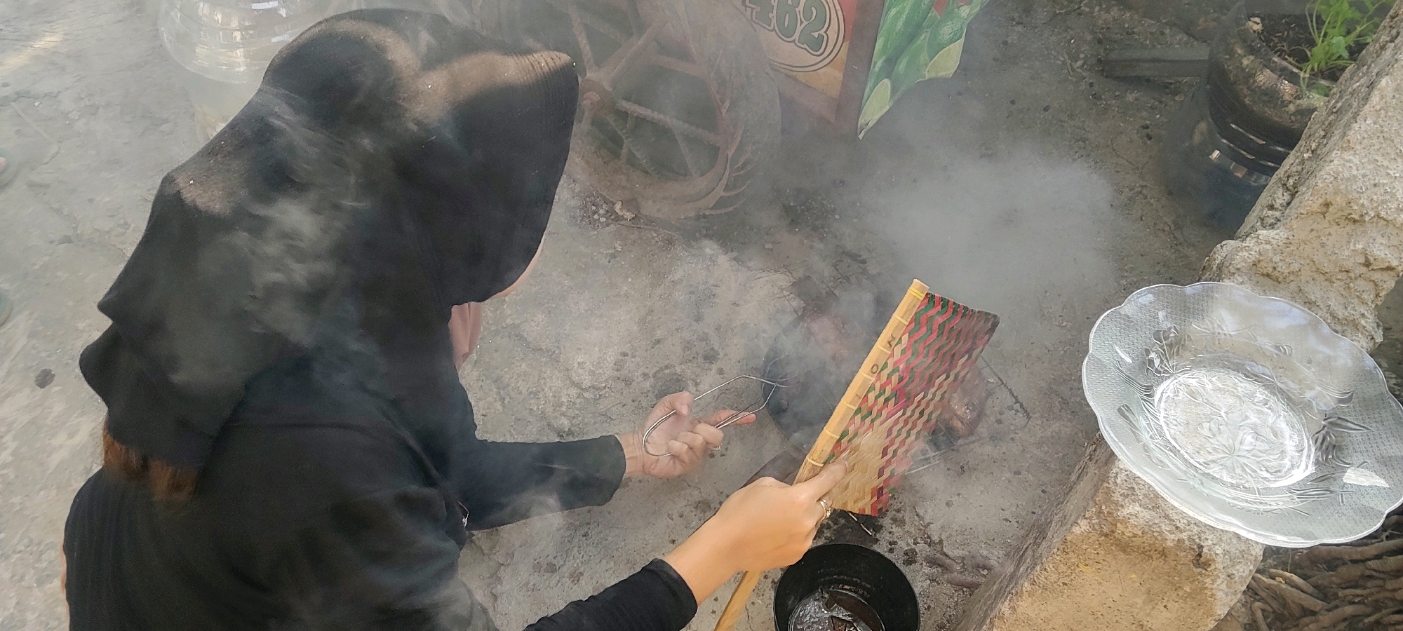 A person wearing a black head covering and a black long-sleeved shirt is seen from behind, fanning a fire with a woven fan. They are using tongs to handle something over the fire, which is producing smoke. In the foreground, a clear glass dish with a floral pattern is visible. The ground appears to be concrete or dirt. In the background, a partially visible water container and some greenery in a pot are present. A rustic sign with text and images is also in the background. The scene suggests an outdoor cooking or preparation activity.