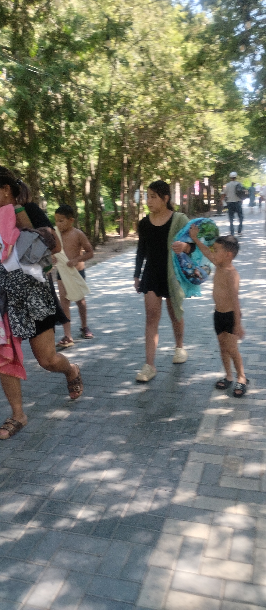 A medium-shot, vertical, blurred photograph shows several people walking on a paved path lined with trees. In the foreground, a person on the left side of the frame, likely female, is wearing a patterned skirt and holding a bundle of clothing. To her right, a young boy is walking, holding a light-colored towel. Further to the right, a young girl in a black dress and a light green shawl is looking to her right. Next to her, a young boy is holding a blue inflatable ring. In the background, other people are visible walking further down the path. The path is made of gray rectangular pavers, and the scene is dappled with sunlight filtering through the trees. The overall impression is that of a sunny day, possibly at a recreational area. The image does not provide specific visual cues to confirm it is Cholpon-Ata, Kyrgyzstan.