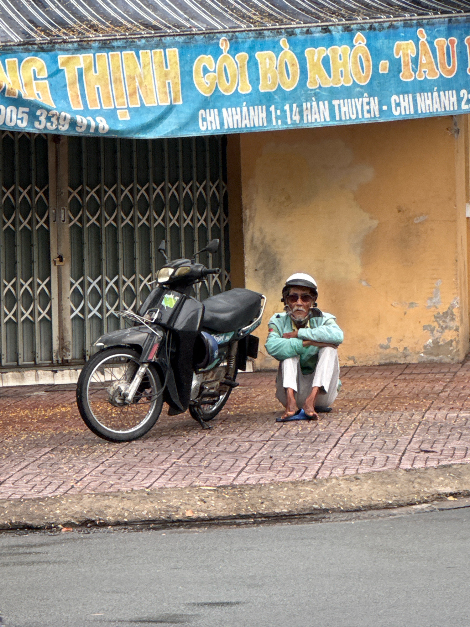 An elderly man wearing a white cap, sunglasses, a light blue long-sleeved shirt, and white pants sits on a brick sidewalk with his arms crossed. He is wearing blue flip-flops. A black motorcycle is parked to his left. Behind them is a yellow building with peeling paint and a blue awning with yellow lettering that reads "NG THỊNH GỎI BÒ KHÔ - TÀU" and "CHÍ NGHÍNH 1: 14 HÀN THUYỀN - CHI NHÁNH 23". The scene is located on a sidewalk in Nha Trang, Vietnam, with a street in the foreground.