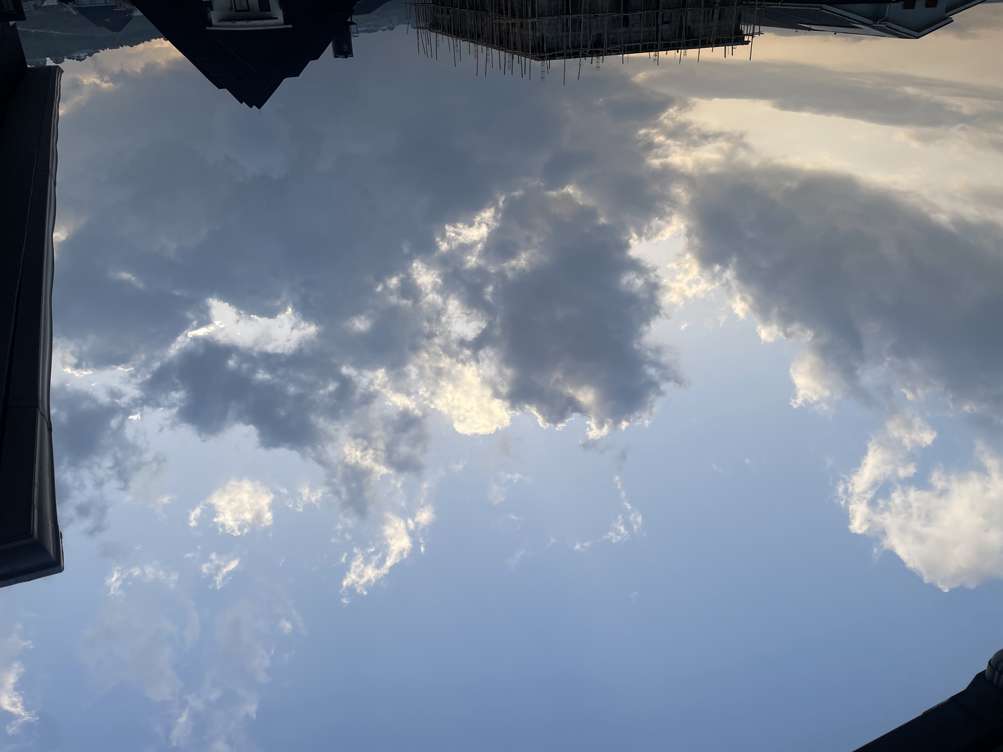 This image shows a cloudy sky over a city, likely Abuja, Nigeria. The sky is a mixture of blue and grey, with sunlight breaking through some of the clouds. Below the sky are buildings, including a partially constructed building with scaffolding and several residential structures with dark, pitched roofs. The foreground on the right is dominated by the edge of a dark building or overhang. The overall scene appears to be taken during daytime, possibly late afternoon or early morning, given the lighting conditions.