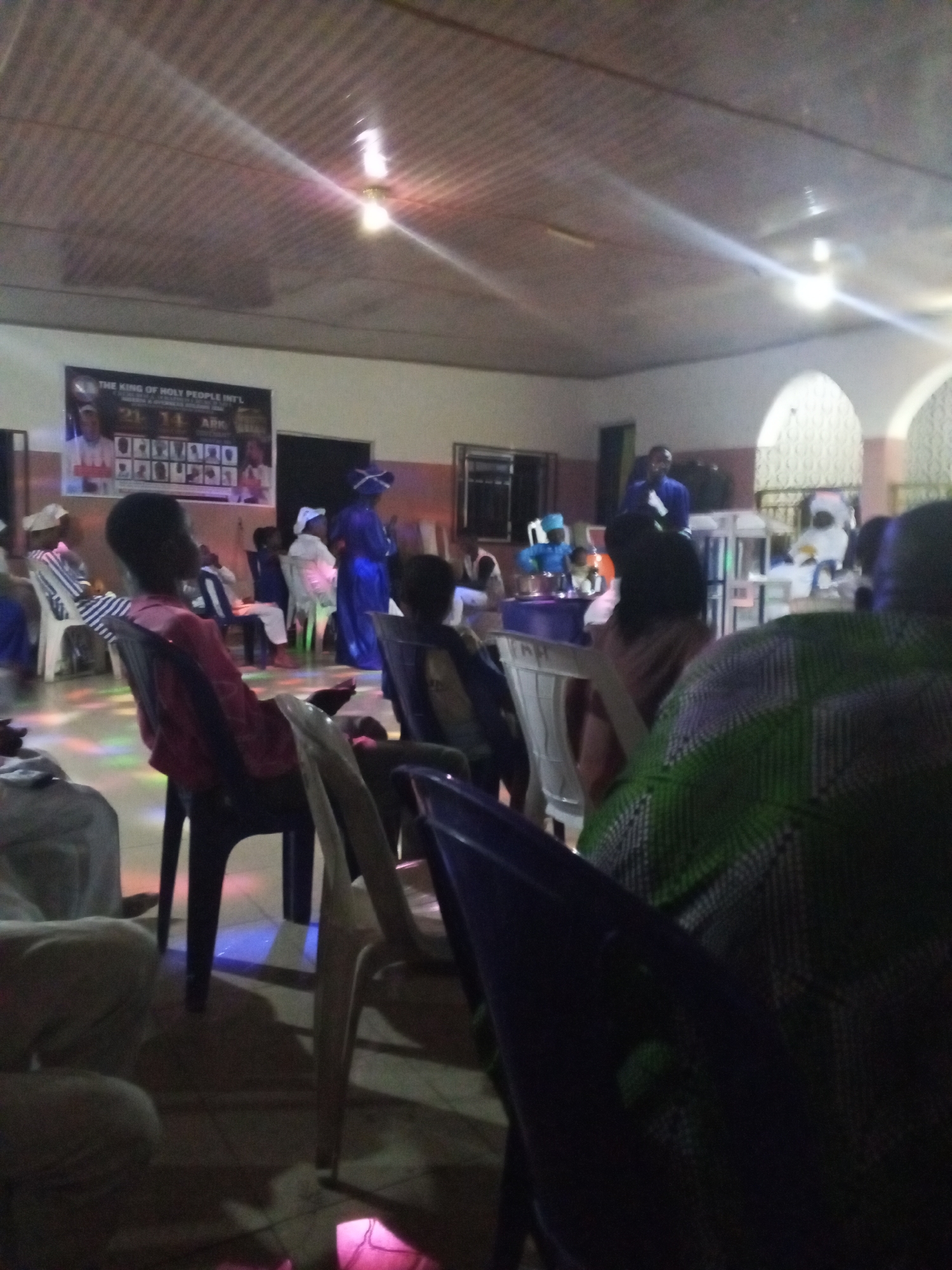 The image shows an indoor gathering in Lokoja, Nigeria. People are seated on plastic chairs. There's a banner on the wall that reads "The King of Holy People Intl". A person in blue robes is standing on a raised platform, possibly addressing the audience. There is a mix of white and blue clothing on many of the people in the room, and the lighting is somewhat dim. Colored light is projected onto the floor.