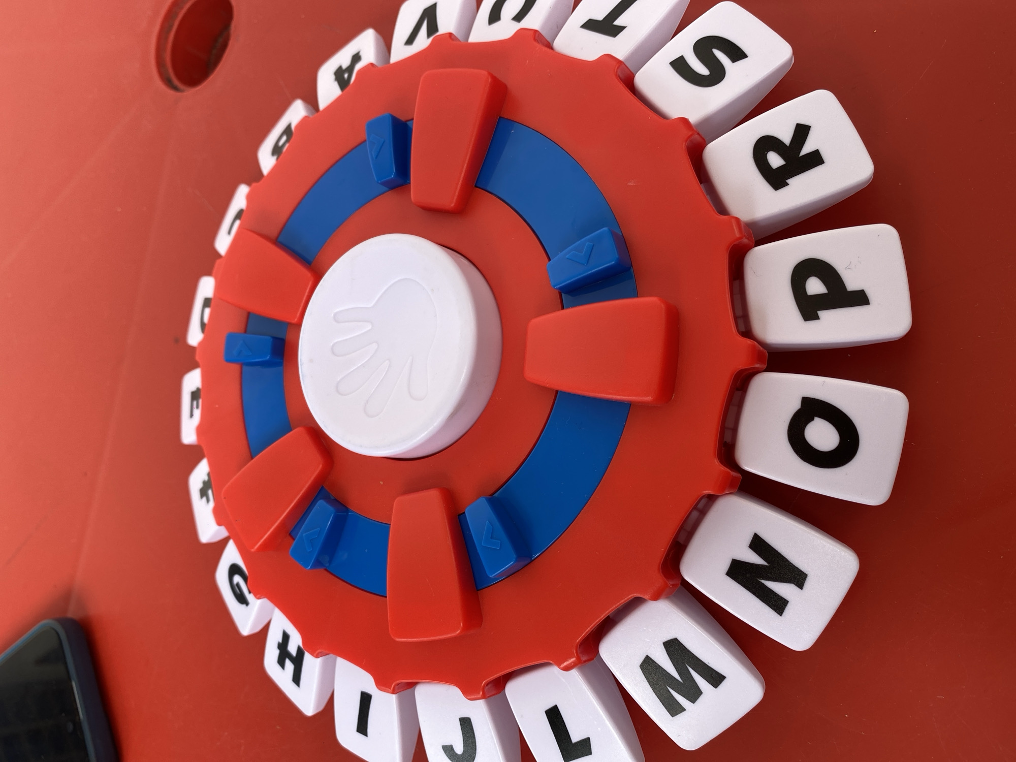 A high-angle shot depicts a red and blue toy with letters arranged around a circular, cog-like perimeter. The letters, printed in black on white tiles, form an alphabet sequence. A white, circular central piece with a hand imprint is surrounded by a blue ring, which in turn is surrounded by red appendages. The entire object rests on a red surface. A partial view of a black phone is visible in the lower left corner. The location is unidentified.