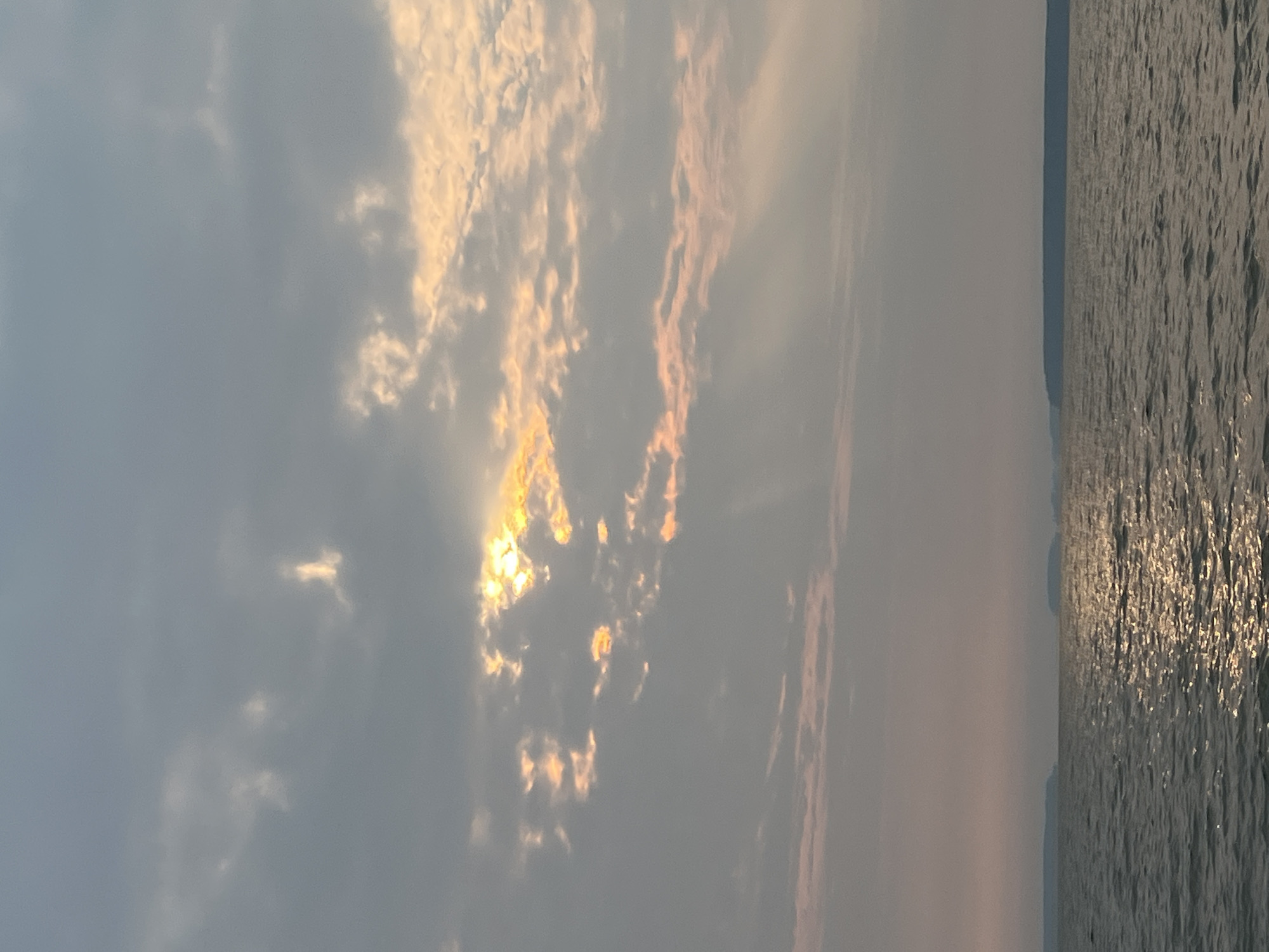 This is a vertical, eye-level, full shot of a body of water with a distant shoreline under a cloudy sky. The water in the foreground is dark with patches of shimmering sunlight reflecting off its surface. The sky is filled with gray and pale pink clouds, with shafts of golden sunlight breaking through in the upper portion of the frame. The distant shoreline is a dark, hazy silhouette of landmasses. The image does not contain any people or specific landmarks that would suggest a location in Novopichugovo, Russia.