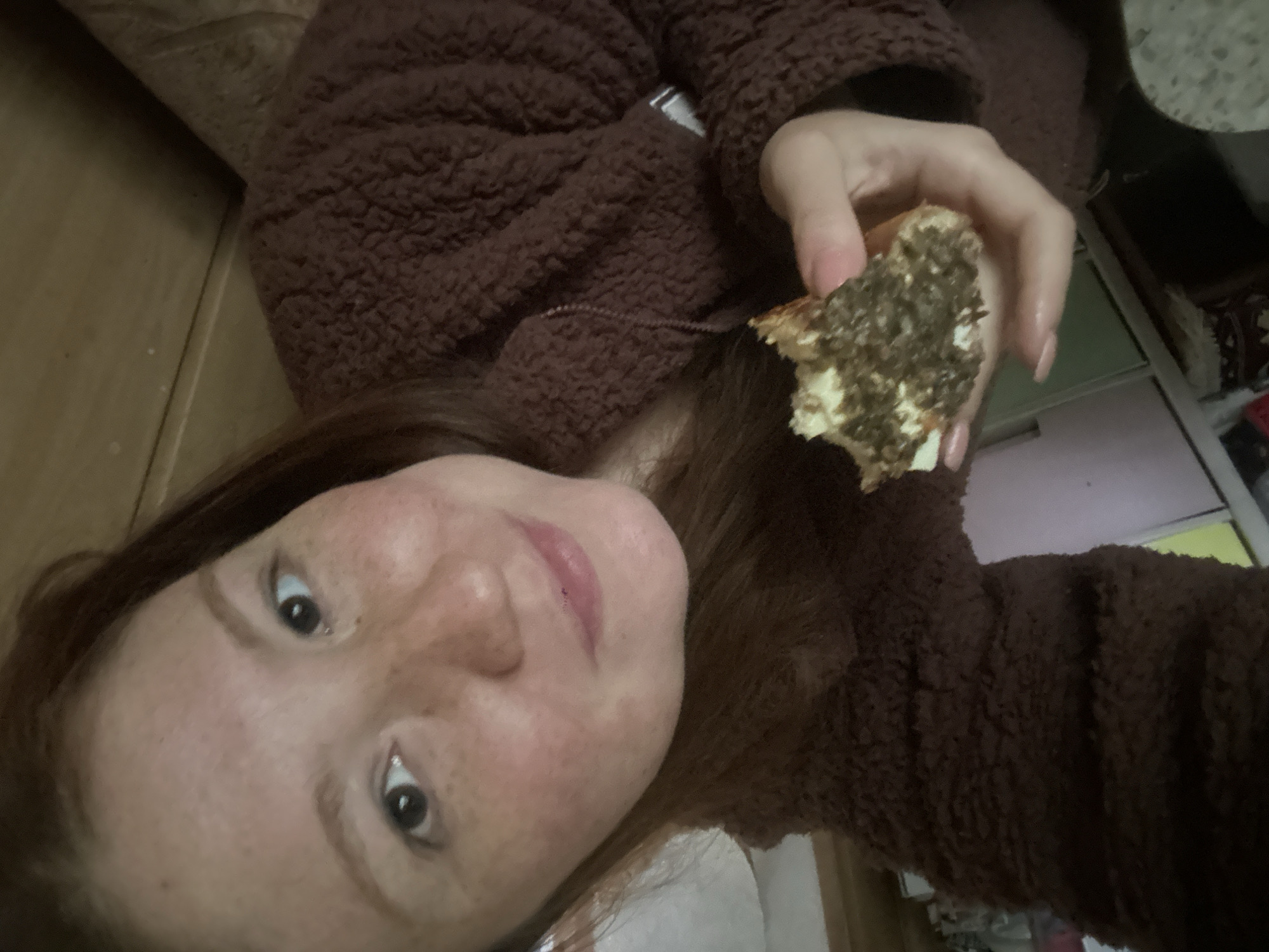 A woman with fair skin, freckles, and auburn hair, wearing a brown fluffy sweater, holds a half-eaten pastry in her right hand. The pastry has a dark topping over a lighter base. The background includes wooden walls, light-colored drawers, and possibly a rug. The location in Nizhny Novgorod, Russia, is not discernible from the image.