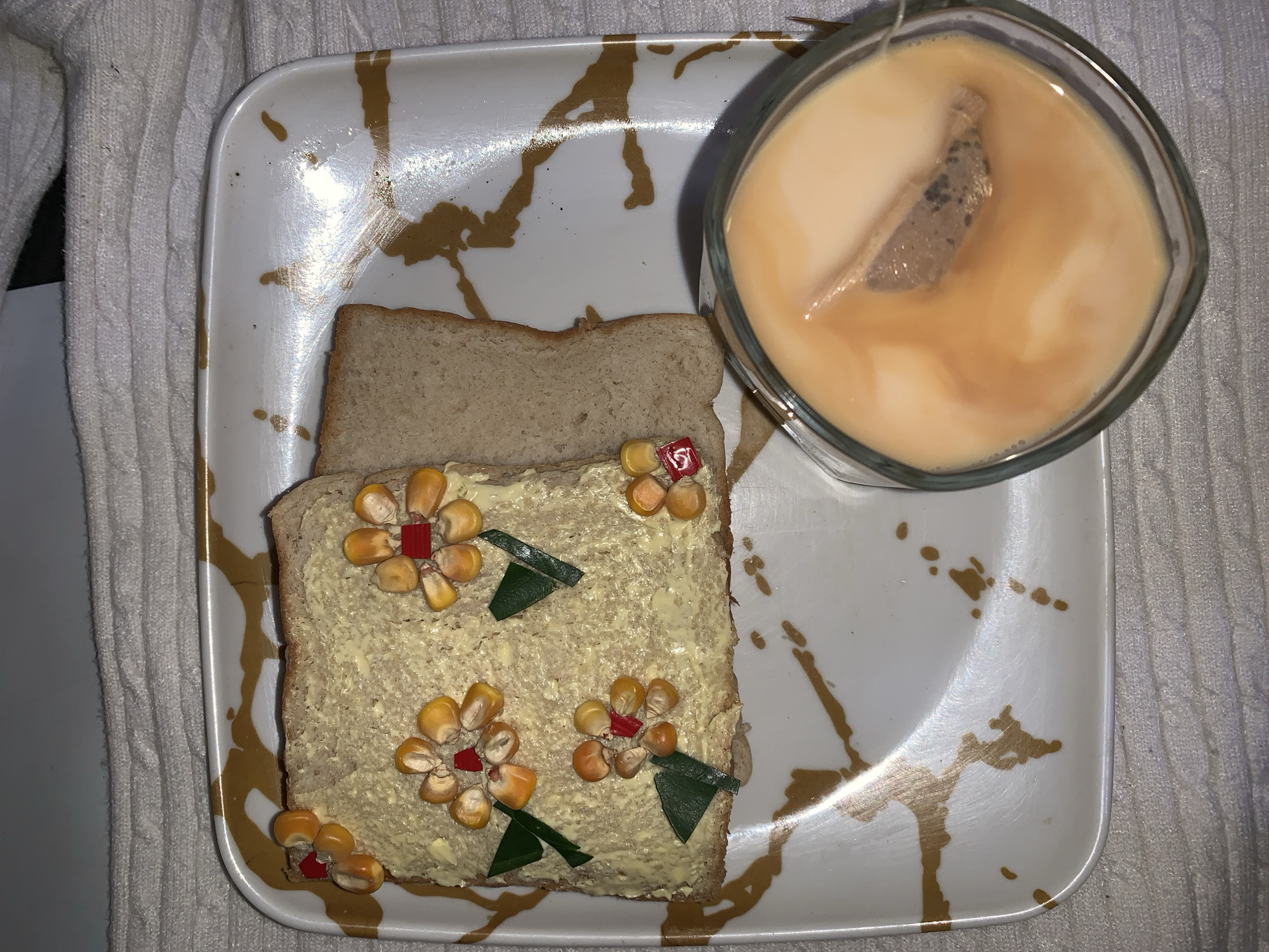 A top-down shot reveals a white square plate with brown marbleized veins. On the plate, there are two pieces of wheat bread, one plain and the other topped with a yellow spread, kernels of corn arranged as flower petals, small red squares at the flower's center, and green leaf-shaped cutouts. Next to the bread is a square glass filled with a milky beige liquid, likely tea, with a tea bag visible inside. The plate is placed on a white textured cloth. The image was taken indoors. There is no evidence this photo was taken in Rafin Par, Nigeria.