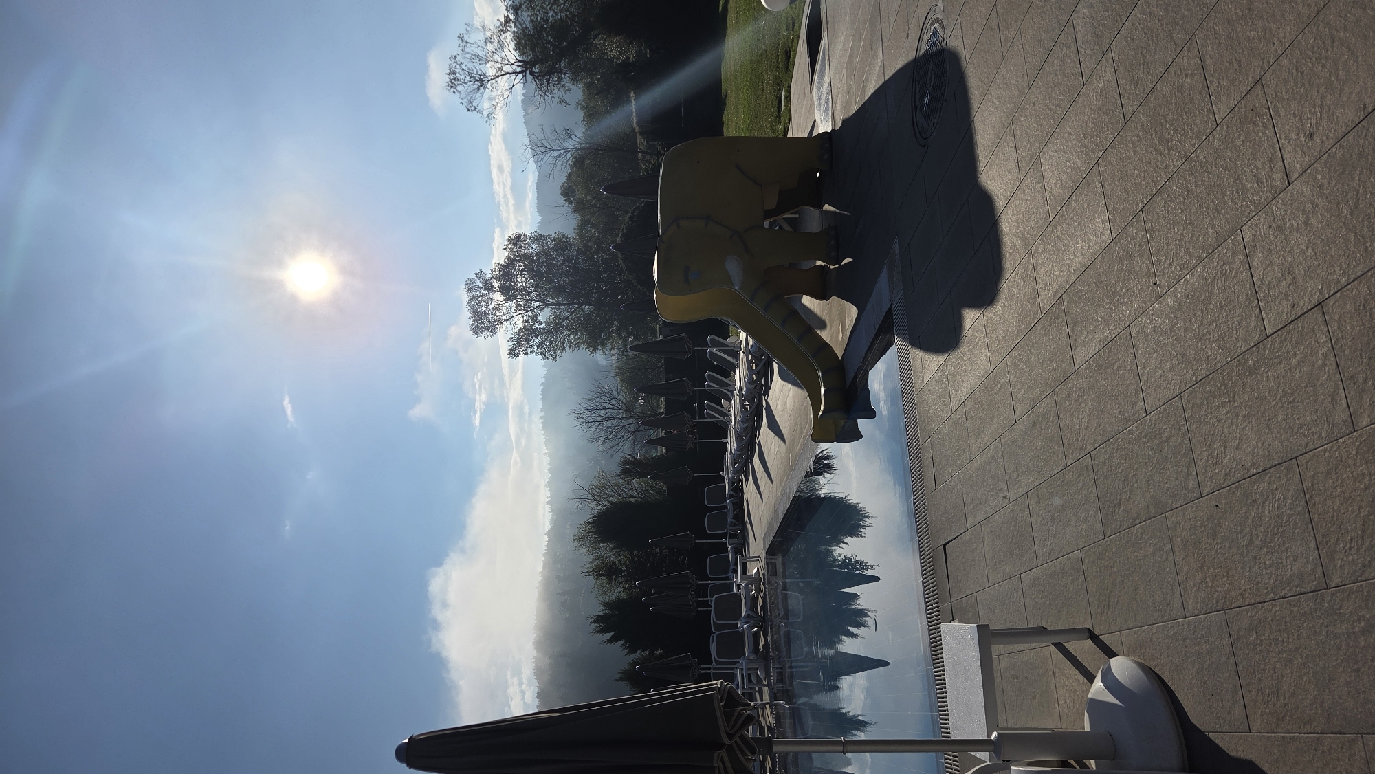 The image depicts an outdoor scene in an unknown location, possibly a resort or hotel. A yellow elephant-shaped structure or slide stands near what appears to be a swimming pool. Lounge chairs and umbrellas are arranged along the poolside. The background features trees and possibly distant mountains shrouded in a hazy atmosphere. The sun shines brightly in the sky with some clouds, suggesting it is daytime. The ground is covered with stone or tile.