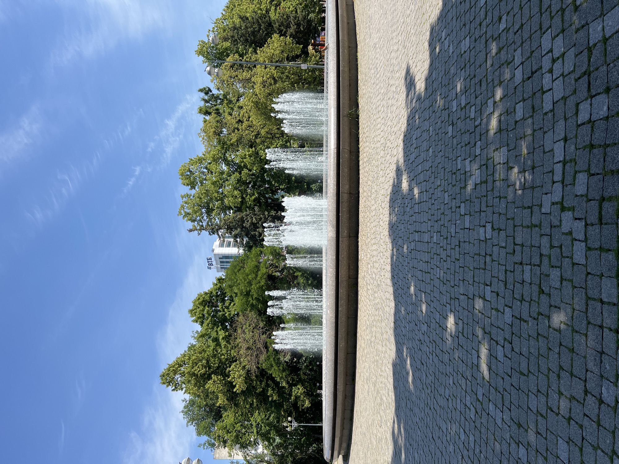 The image depicts a wide, circular fountain in a paved public square. The fountain has multiple jets of water spraying upwards, forming a shimmering curtain. A thick, polished stone rim encircles the fountain. The paved area in front of the fountain is made of small, gray cobblestones, with shadows of trees cast upon it. Behind the fountain, a dense line of lush green trees forms a backdrop. In the distance, above the trees, a modern white building with the blue Cyrillic letters "ВТБ" is visible. The sky above is a clear, bright blue with a few wispy white clouds. A streetlamp is visible to the left of the fountain, and another is partially visible to the right. No people are clearly visible in the immediate vicinity of the fountain, though a person can be seen briefly on the far right, near the fountain's edge, possibly tending to a plant or interacting with the fountain's edge. The scene appears to be set in a park or city square on a sunny day. The presence of the "ВТБ" building suggests the location is likely in a city where this bank has a significant presence, such as Krasnodar, Russia, given the Cyrillic script.