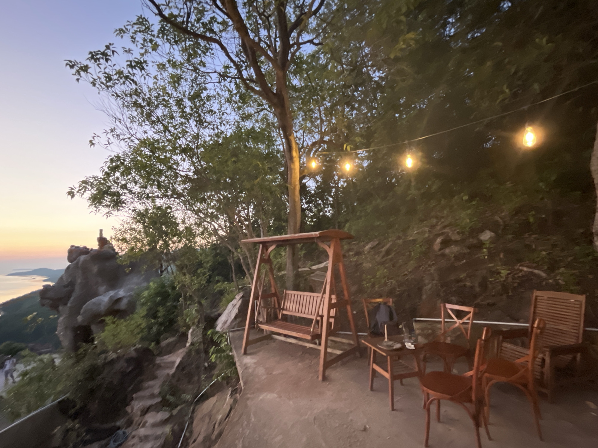 This is a photograph of an outdoor seating area with a view of the ocean at dusk. In the foreground, there is a wooden swing set, a small wooden table, and several wooden chairs. The area appears to be on a cliff overlooking the sea, with a path leading down the rocks on the left. String lights are visible hanging from trees, casting a warm glow on the scene. The sky is a gradient of pale blue to soft orange, indicating either sunrise or sunset. The background is filled with lush green trees and foliage. The location is identified as Karimunjawa, Indonesia.