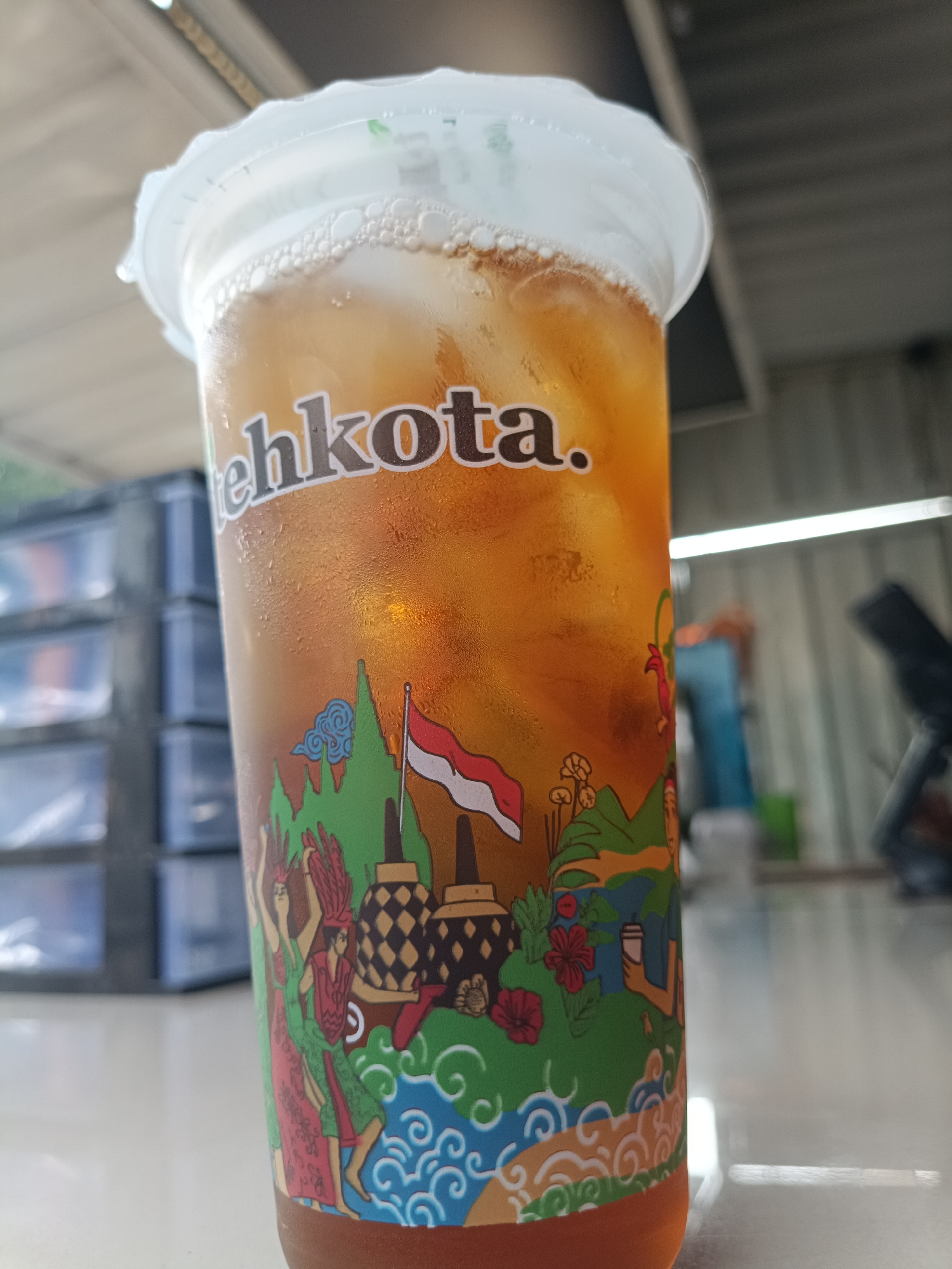 A close-up, eye-level, medium shot shows a clear plastic cup filled with iced tea. The cup has a white plastic lid and is condensation-covered. A decorative band wraps around the lower half of the cup, featuring a colorful, cartoonish depiction of Indonesian cultural elements, including a flag, traditional buildings, dancers, mountains, and water motifs. The brand name "tehkota." is visible in white lettering on the upper part of the cup. The cup rests on a white surface, possibly a table, with a blurred background that includes stacked dark blue drawers on the left and a bright fluorescent light running horizontally across a metallic wall on the right. The setting appears to be an outdoor or semi-outdoor food stall or eatery. The location is identified as Jember, Indonesia.