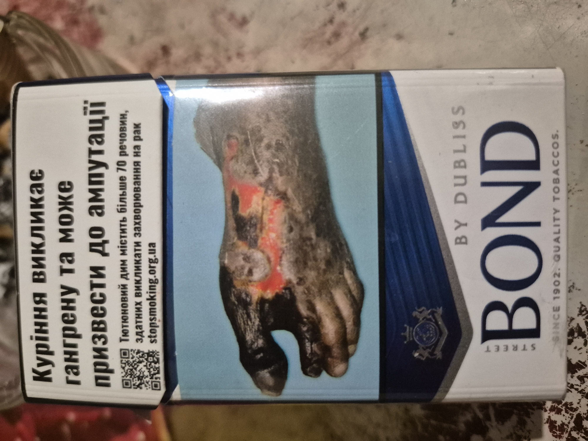 This is a close-up, top-down shot of an open pack of Bond cigarettes, featuring a stark health warning with a graphic image of a diseased hand. The packaging is predominantly white with blue accents and a large, bold "BOND" logo. The health warning in Ukrainian text states that smoking causes gangrene and can lead to amputation, also mentioning that tobacco smoke contains over 70 carcinogens and causes cancer. A QR code is present, linking to stopsmoking.org.ua. The background is blurred, suggesting the pack is resting on a surface, possibly a table or counter. No specific location in the city of Marhanets, Ukraine is discernible from the image itself.