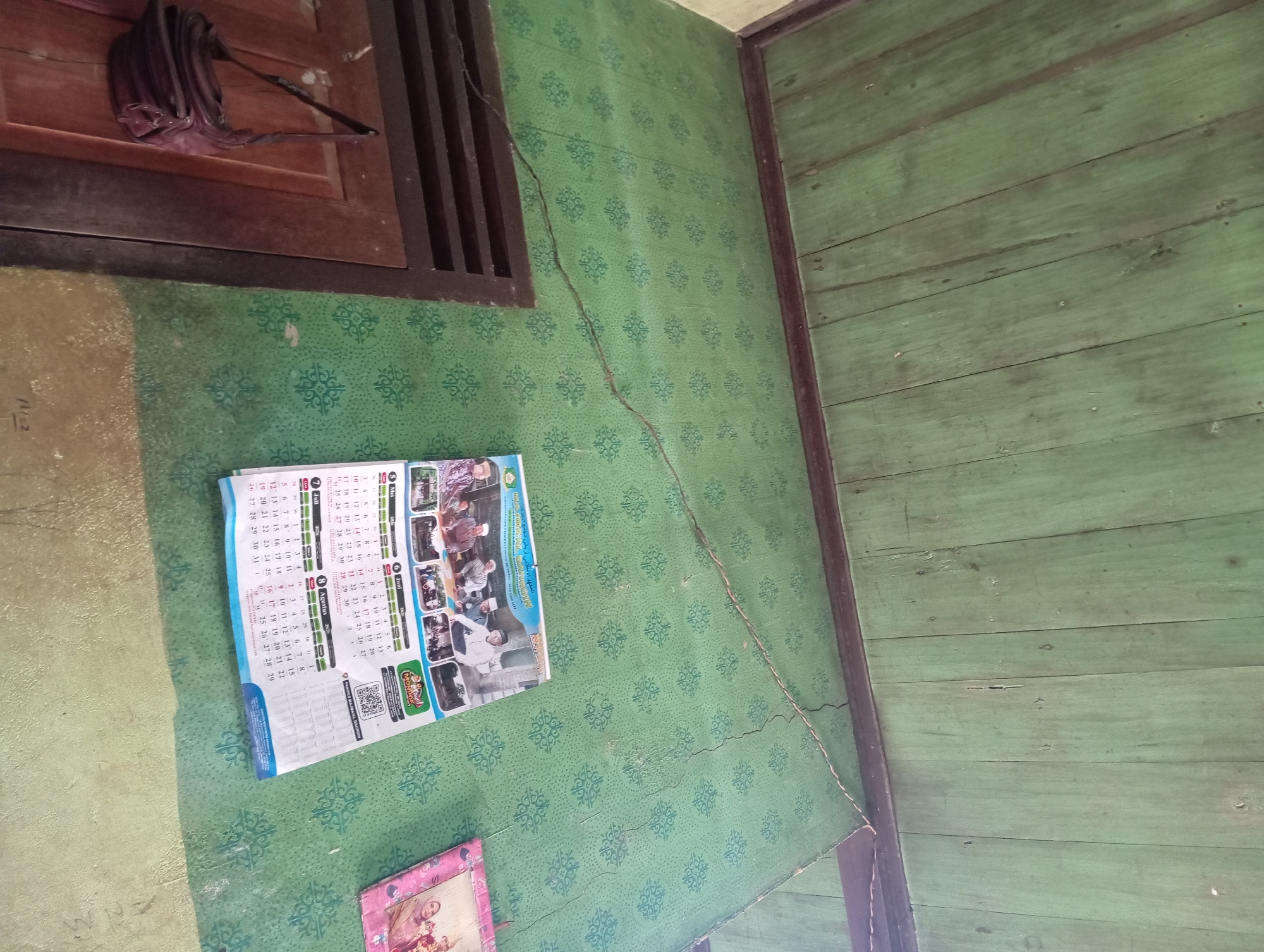 The image shows an interior view of a room in Rejosari, Indonesia. The walls are covered with green patterned wallpaper, and the ceiling is made of green-painted wooden planks. A brown wooden door is visible on the left side of the image, with a dark red handbag hanging from it. A calendar is displayed on the wall, featuring a group of men in religious attire and a QR code. A framed picture is also present on the wall to the right of the calendar. The room appears to be in a state of disrepair, with cracks visible on the wall and ceiling. The lighting suggests it is daytime, but the exact time is not discernible from the image.