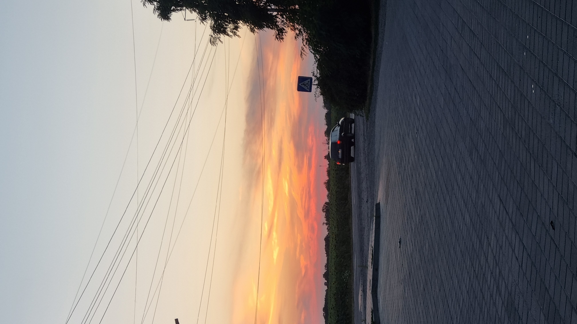 The image captures a sunset over a rural landscape near Smila, Ukraine. A dark-colored car is driving on a paved road in the foreground, with a pedestrian crossing sign visible on the right side of the road. The sky is illuminated with vibrant orange and pink hues, indicating the sun is low on the horizon. Fields of crops, possibly sunflowers, stretch across the background, bordered by trees. Power lines crisscross the sky, partially obscuring the sunset. The foreground pavement is made of interlocking rectangular blocks. The scene appears to be an intersection or a roadside area on the outskirts of a town or village.