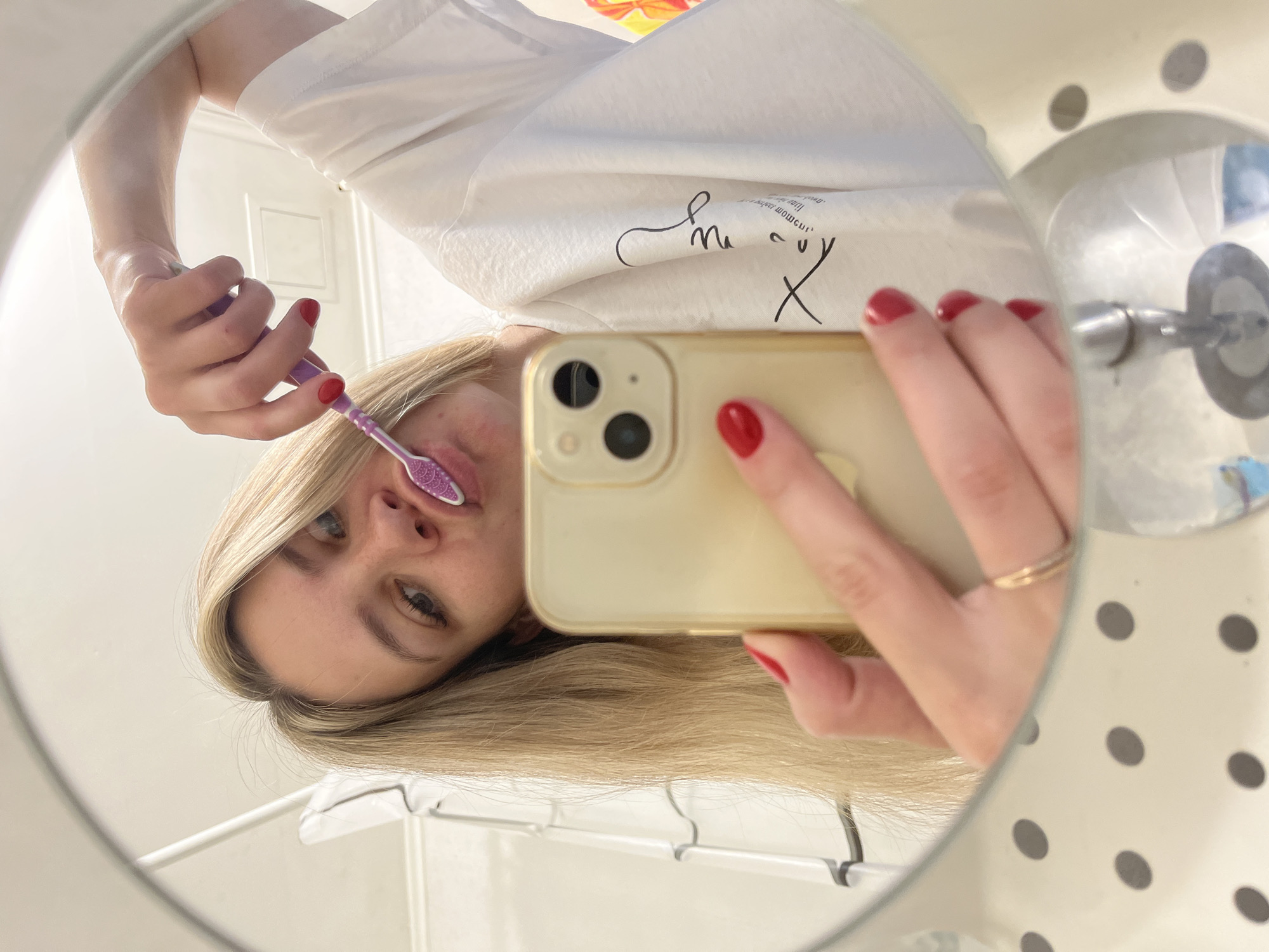 A woman is brushing her teeth in a bathroom mirror selfie. She has blonde hair and is wearing a white t-shirt with a graphic design on it. She is holding a pink toothbrush in her right hand and her phone in her left hand. Her nails are painted red. The bathroom appears to be modern, with white tiled walls and a speckled countertop. The image is taken from a low angle, reflecting the woman and her surroundings in the mirror. The scene is consistent with a personal bathroom in a residential setting, and there is no specific information to suggest it is located in the city of Chaikovsky, Russia.