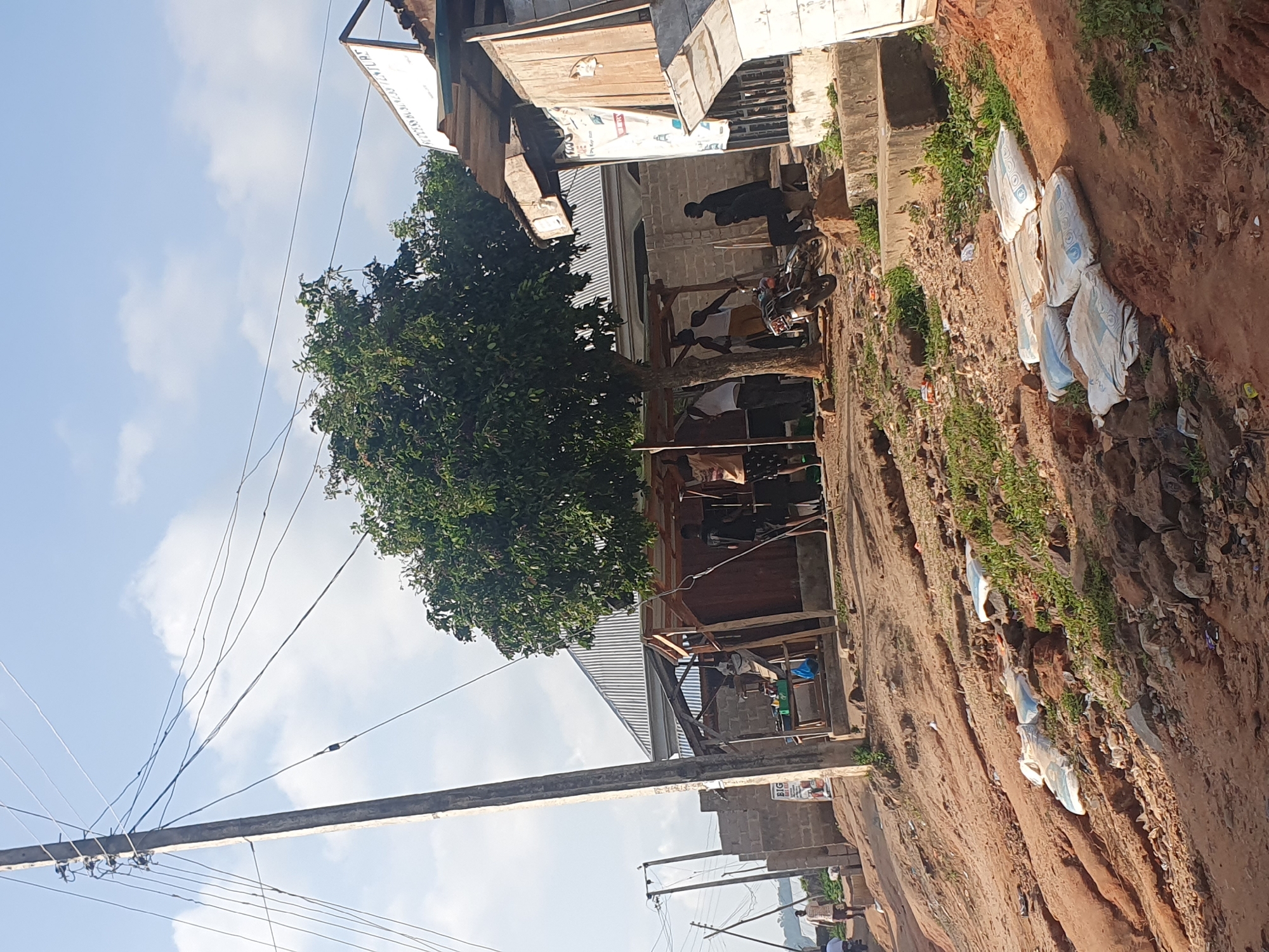 The image depicts an outdoor scene, possibly in Lokoja, Nigeria, under a bright, cloudy sky, suggesting daytime. The foreground is dominated by a dirt road and a large tree with lush green foliage. To the left, a wooden utility pole supports a network of power lines, some of which crisscross the frame.
Beyond the tree, a collection of structures, appearing to be shops or informal dwellings, line the road. These buildings are constructed from various materials, including corrugated metal roofing and wooden planks. In front of one of these structures, several people are gathered, some standing and others seated. A motorcycle is parked nearby, and sacks are visible, some stacked along the roadside, others possibly containing goods. The general atmosphere appears to be that of a busy, possibly commercial, area.
There is some visible text on a white banner hanging from one of the buildings, which reads "BIG" in large letters, followed by smaller, less legible text. The scene suggests a rural or semi-urban setting with everyday activities taking place. The orientation of the image is rotated 90 degrees counter-clockwise.