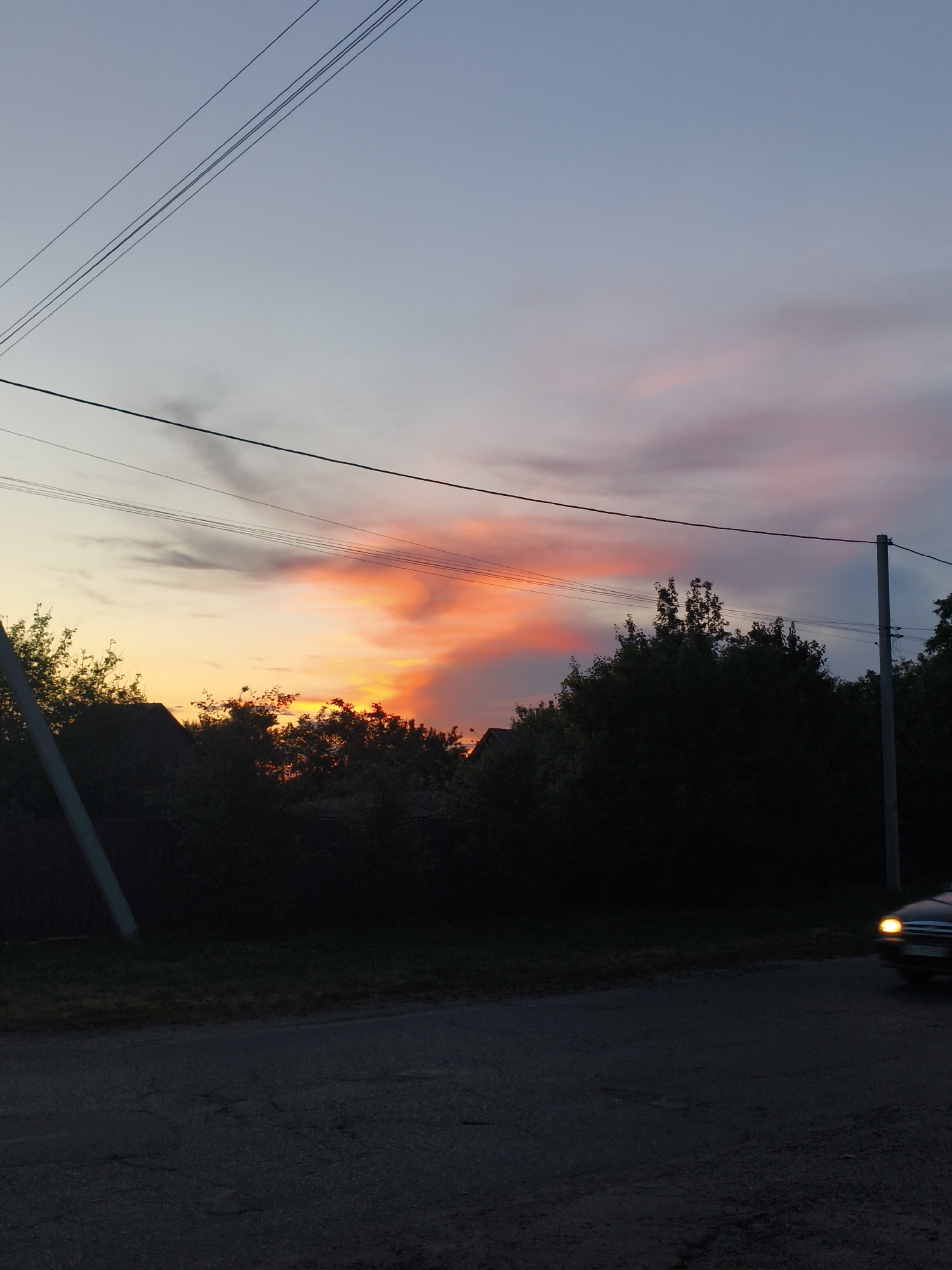 The image captures a rural twilight scene in Ustyvytsia, Ukraine. The sky is a gradient of pale blue at the top, transitioning to soft pink and fiery orange where the sun has set, illuminating the clouds. Silhouetted against this vibrant sky are the dark shapes of trees and the roofs of houses. Power lines crisscross the upper portion of the frame, supported by utility poles. A dark road runs through the foreground, with a dark-colored car visible in the lower right, its headlight on. The overall atmosphere is serene and tranquil, depicting the quiet beauty of a rural evening.