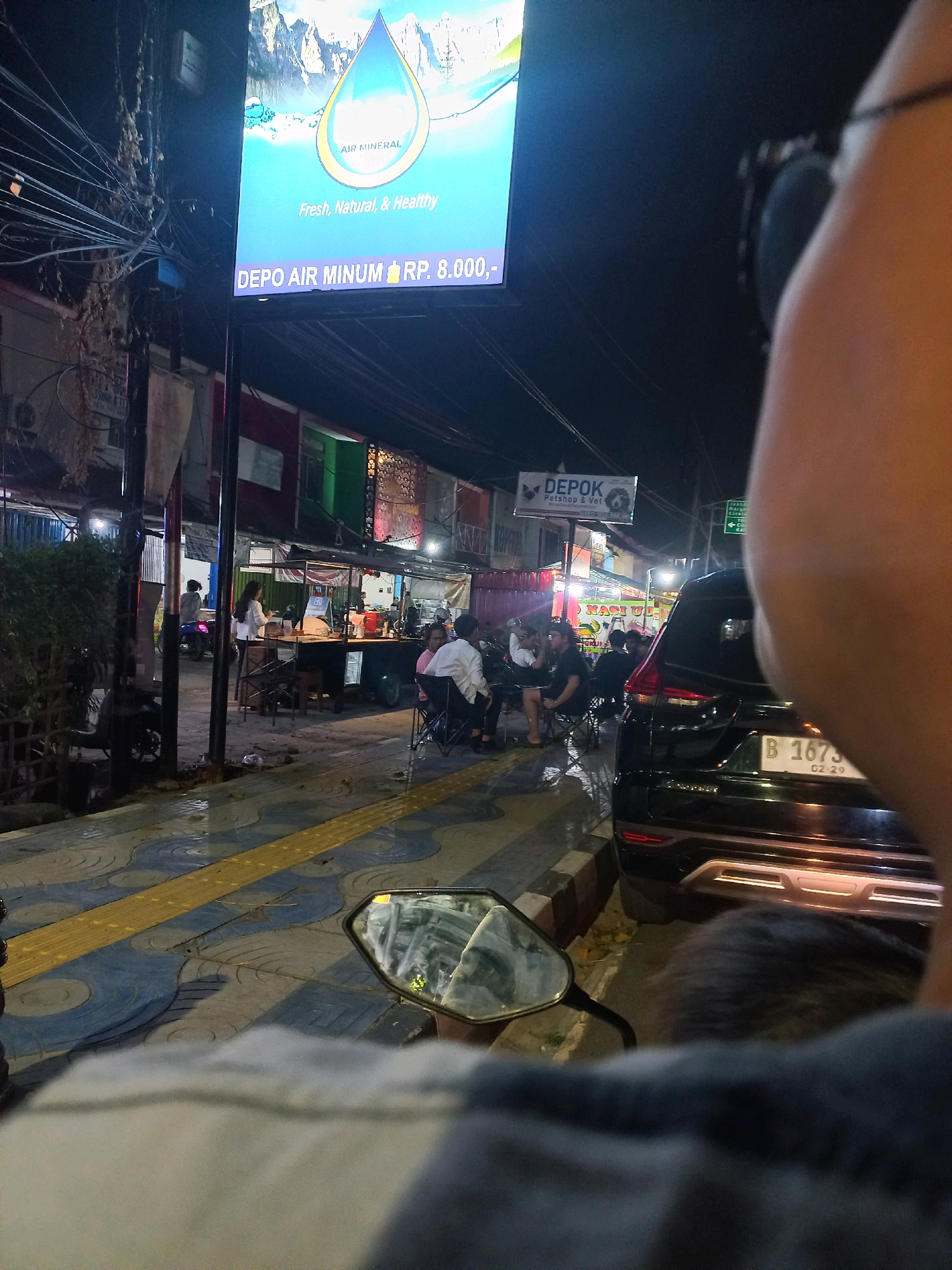 The image depicts a street scene at night in Depok, Indonesia, near a "Depo Air Minum" (drinking water depot). A large illuminated sign above the depot advertises "AIR MINERAL Fresh, Natural, & Healthy" and displays the price "RP. 8.000,-". In the foreground, a portion of a person's head and shoulder is visible, suggesting the photo is taken from the perspective of someone on a motorcycle. A motorcycle rearview mirror reflects the scene, including a glimpse of the rider's head and the street. The street is paved with decorative tiles and a yellow tactile paving strip, indicating a pedestrian area. Several people are seated at tables outside a food stall or small restaurant on the right side of the street, with some appearing to be eating or conversing. A black SUV is parked on the street, its license plate visible as "B 1673 QZ". A smaller sign for "Depok Petshop & Vet" is also visible further down the street. The environment appears to be urban, with electrical wires crisscrossing overhead and building facades lining the street. The lighting suggests nighttime, with the main illumination coming from the signs and streetlights.