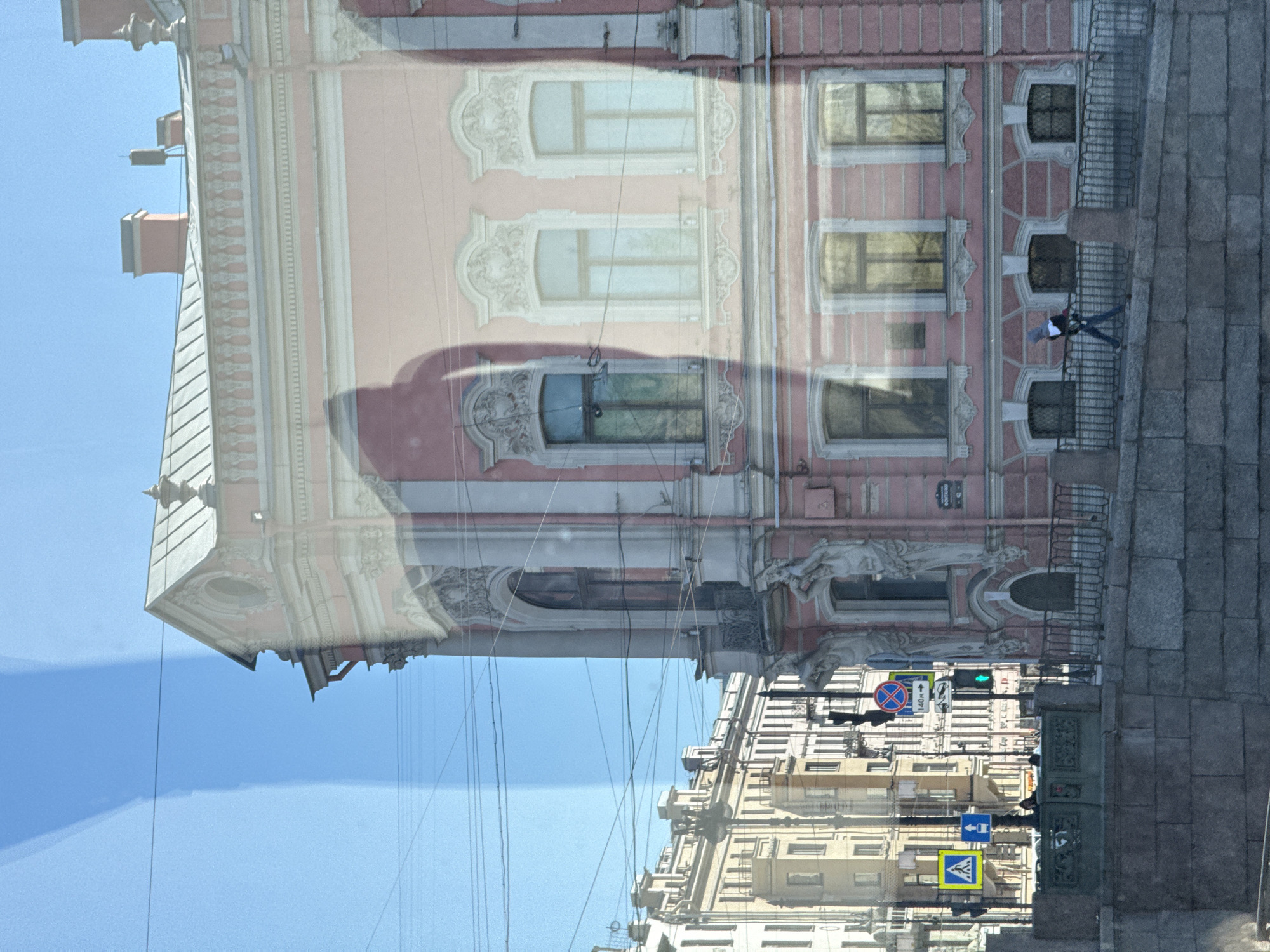 The image shows a reflection of a building in Saint Petersburg, Russia, in a window. The building is light pink with white trim and ornate window detailing. A pedestrian is standing on a stone walkway in front of the building, behind a metal railing. The reflection shows multiple other buildings, power lines, and street signs. The sky is blue.