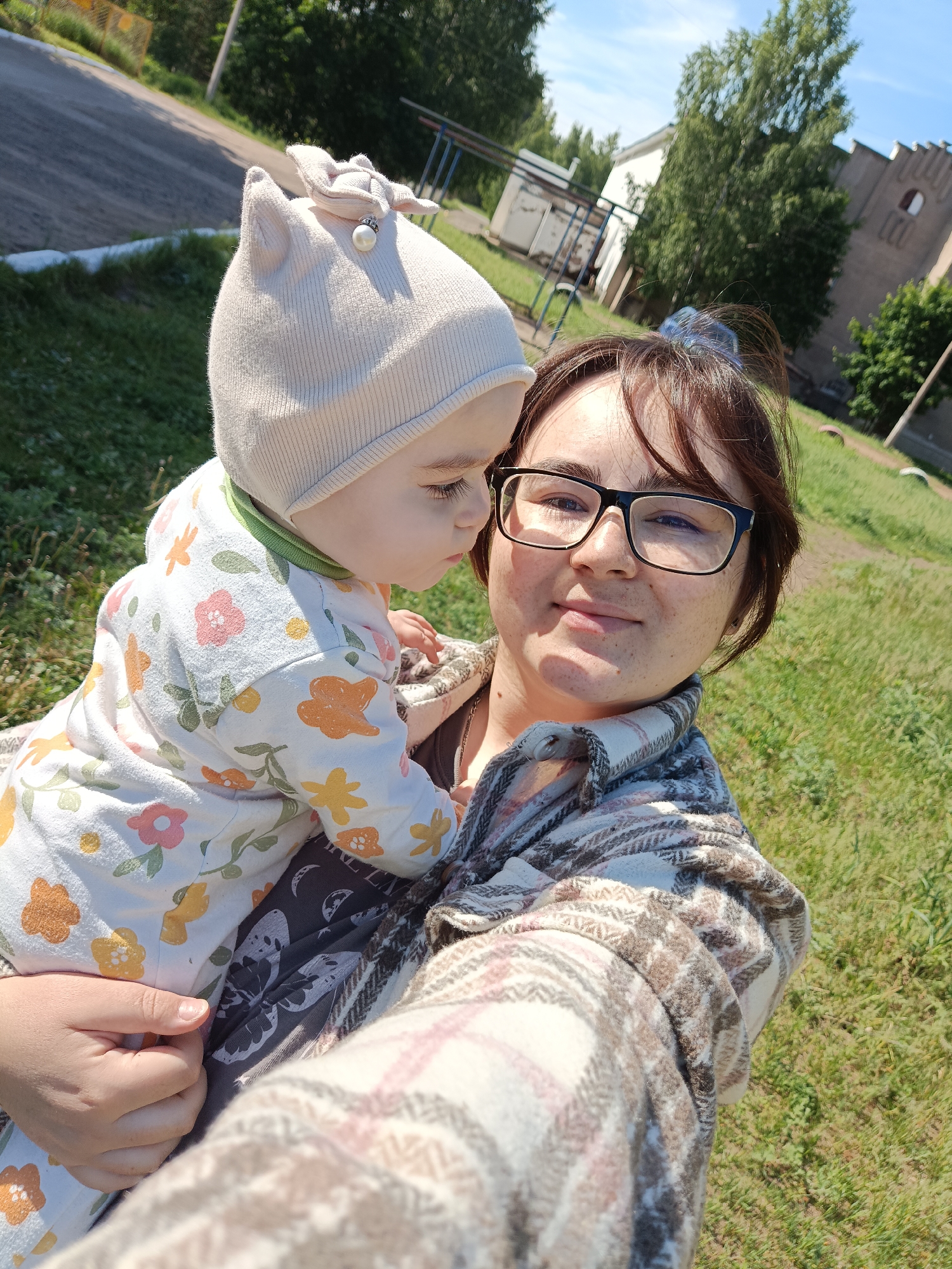 A woman and a baby are featured in a sunlit outdoor selfie, likely in Noviy, Russia. The woman, with dark hair tied back and wearing black-rimmed glasses, a plaid flannel shirt (beige, brown, pink), and a dark top, smiles gently at the camera. She has visible freckles on her face. She holds the baby, who is dressed in a light-colored onesie adorned with a floral pattern of pink, orange, and green flowers and leaves. The baby also wears a light beige beanie with subtle cat ears and a decorative bow featuring a pearl and small rhinestones, and is looking towards the woman's face.
The scene is set outdoors on a bright, clear day, likely morning or afternoon, given the strong sunlight and shadows. In the background, there's a grassy area, an asphalt road or path, and some simple metal playground equipment. Further back, buildings and trees are visible. One building appears to be light-colored, while another to the right has a distinct, possibly older, facade with arched architectural elements, suggesting a village or small town environment. The overall mood is warm and affectionate. There is no visible text in the image.