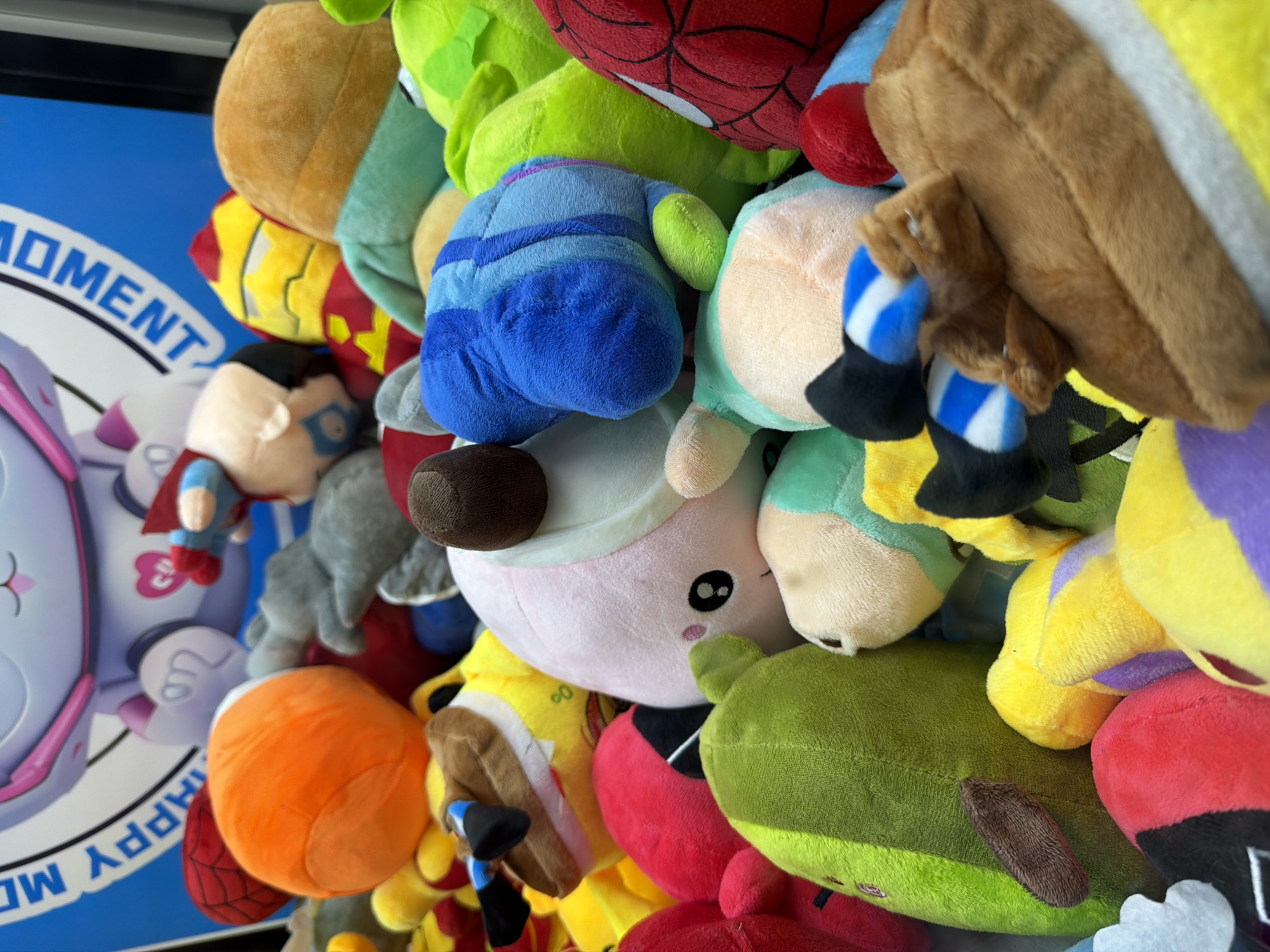 The image presents a vibrant, close-up view of a large, densely packed collection of soft toy plushies. The toys are colorful and varied, suggesting they are part of a claw machine, prize display, or retail bin. Recognizable characters include a red Spider-Man with black web detailing, a red and yellow Iron Man, and a blue Superman with a red cape. Other notable plushies feature a squat blue character, possibly from Among Us; a light pink character with black eyes and rosy cheeks; a green block-shaped toy, potentially a Minecraft Creeper; and yellow characters, one appearing bee-themed.
The plushies are stacked haphazardly, filling the majority of the frame and creating an impression of abundance. In the background, a bright blue graphic is partially visible. This graphic features a large, light-colored character, possibly a stylized cat or robot, adorned with pink accents and a heart symbol on its chest. Fragments of text are discernible on this background, including "OMENT" (likely part of "MOMENT") and "HAPPY MOW" (possibly "HAPPY MOMENT" or "HAPPY NOW"), suggesting a cheerful or celebratory theme. The overall scene implies an indoor setting, such as an arcade, entertainment center, or toy store in Sliven, Bulgaria. The lighting is consistent with an interior environment. No specific activity is depicted other than the arrangement of the toys.