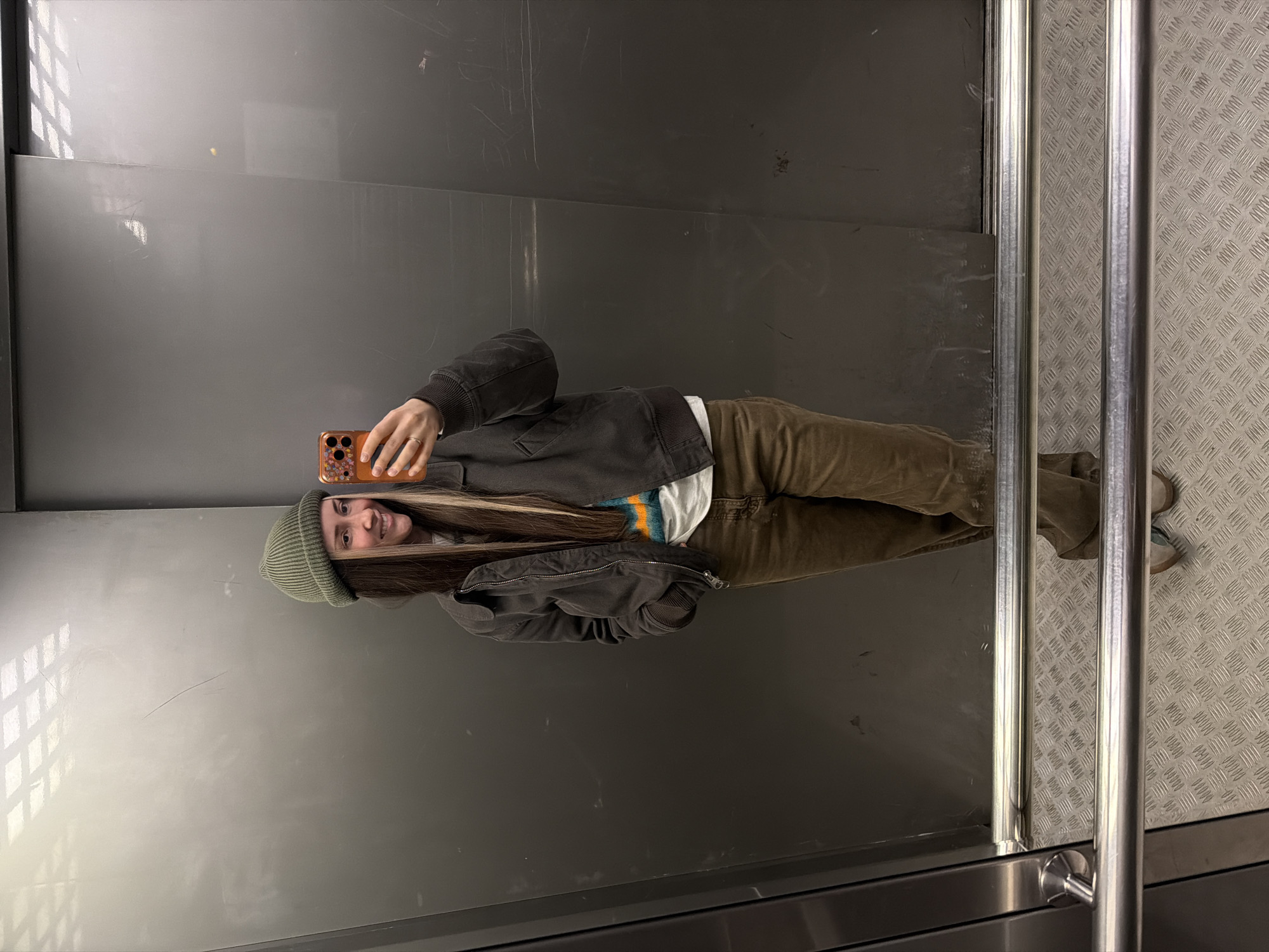 A person wearing a green beanie, brown jacket, and brown pants is taking a selfie in what appears to be an elevator or enclosed space. The background is gray and metallic, with vertical silver bars visible on the right side. The floor has a textured pattern. No specific details indicate that the location is Khimki, Russia.