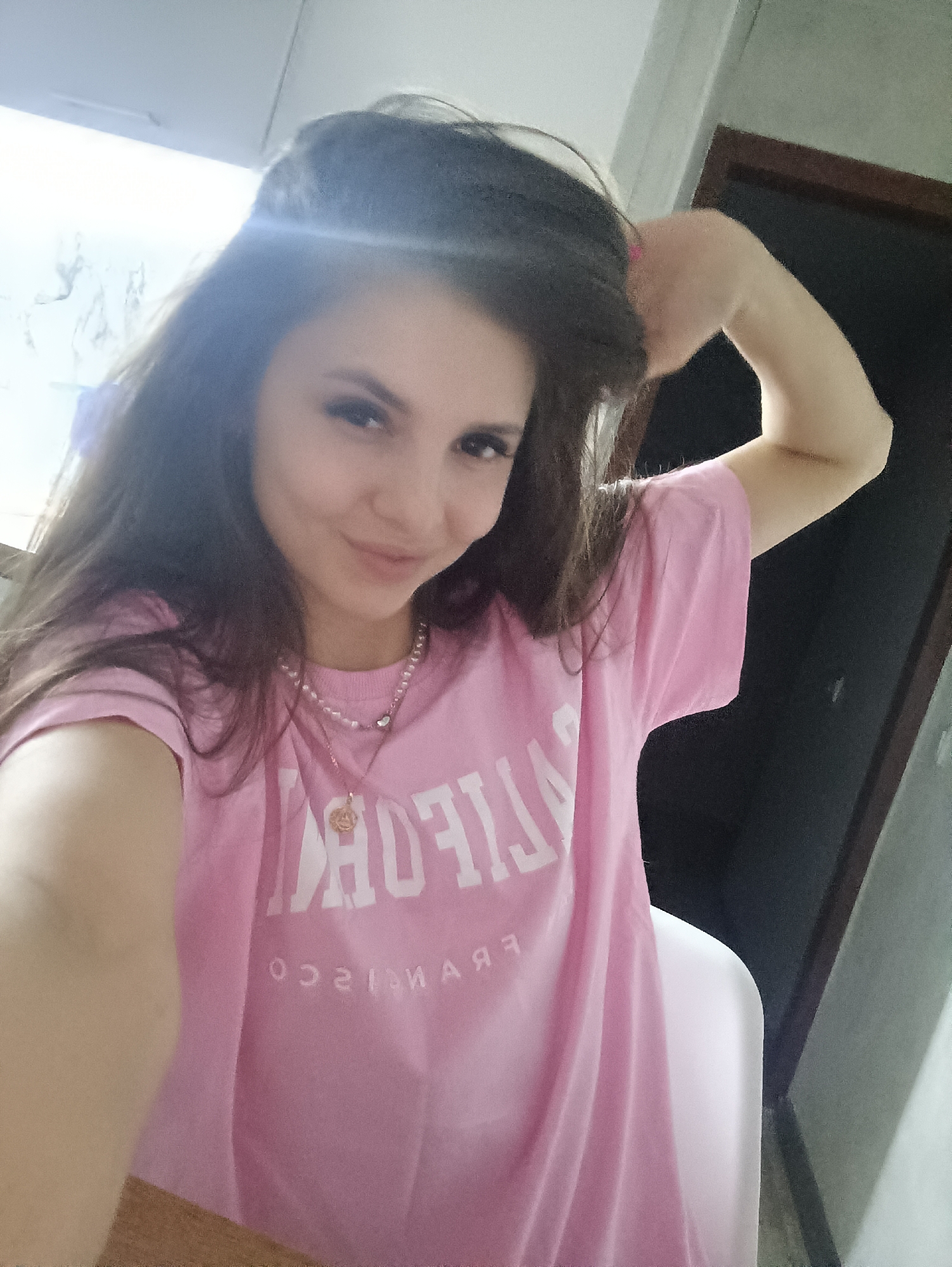 A young woman with long, dark brown hair is posing for a selfie. She is wearing a pink t-shirt with the words "CALIFORNIA" and "SAN FRANCISCO" printed on it in white letters. She is also wearing a pearl necklace with a gold pendant. Her right arm is raised and her hand is touching her hair. The background is blurred, but it appears to be an interior space with a door and some cabinetry. The location is unknown and there is nothing to indicate that this is in the city of Chaikovsky, Russia.