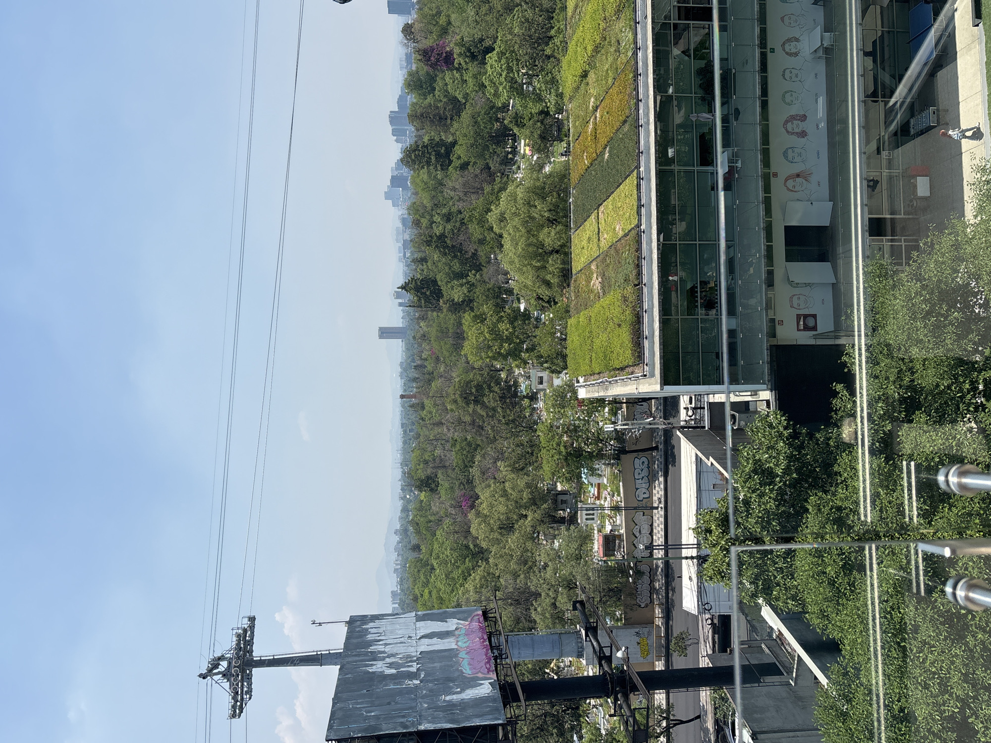 The image depicts a view from a high vantage point in Mexico City, Mexico. The foreground features modern architecture with a green roof and reflective glass walls adorned with images of faces. Adjacent to these buildings are older structures displaying graffiti and electrical utility infrastructure. A cable car support tower and associated lines dominate the left side of the frame. In the distance, a dense canopy of trees blankets the landscape, punctuated by the silhouettes of city buildings against a hazy, light blue sky.