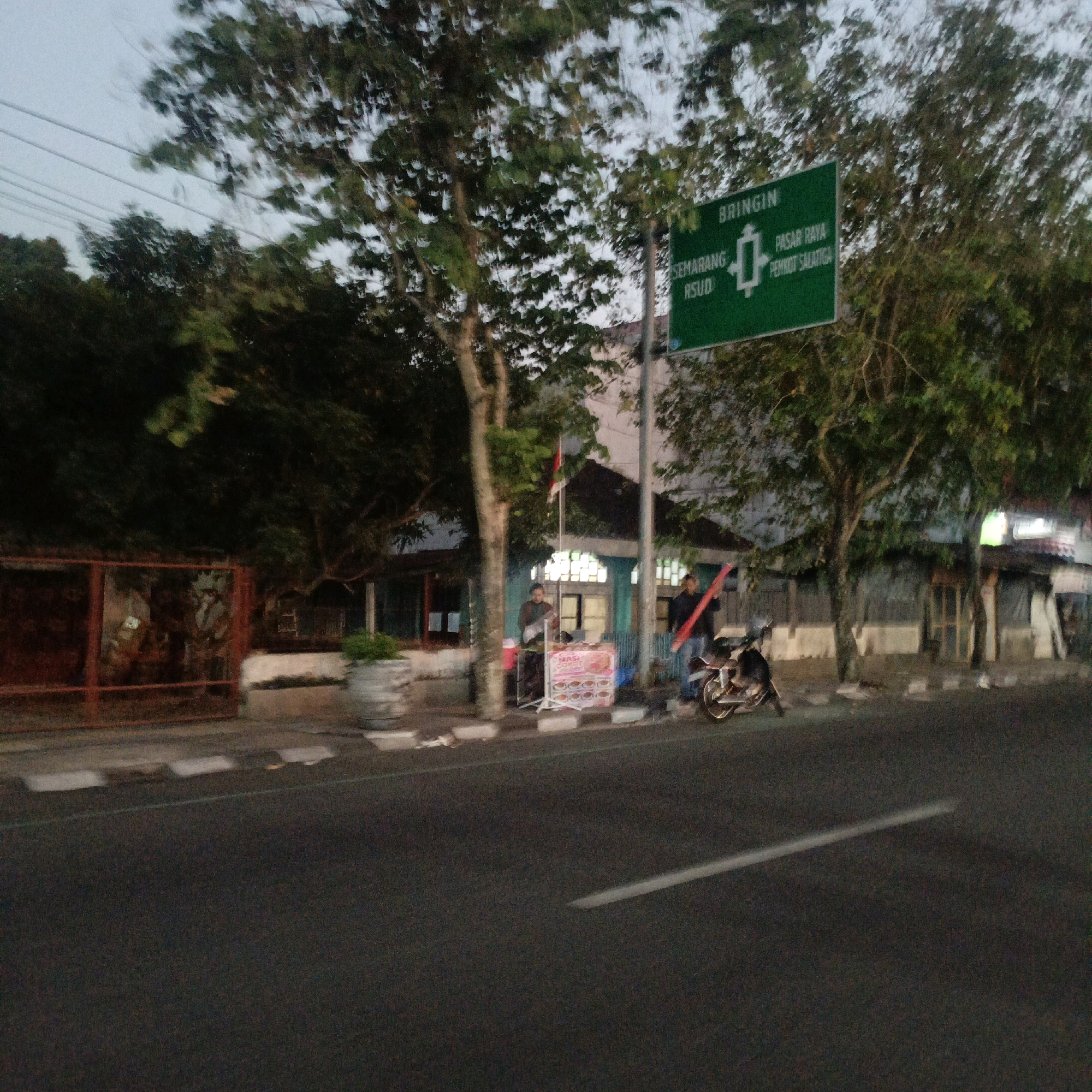 The image depicts a street scene in Salatiga, Indonesia, near a building with a green directional sign. The sign indicates "BRINGIN" at the top, with arrows pointing left to "SEMARANG RSUD" and right to "PASAR RAYA PEMKOT SALATIGA". Two individuals are visible on the sidewalk near a food stall. One person, wearing dark clothing, stands behind the stall which displays images of food. Another person, in blue jeans and a dark top, holds a red object. A motorcycle is parked on the side of the road. Trees line the street, and a fence is visible on the left side of the image. The sky is overcast, suggesting it might be early morning or late afternoon. The road surface is asphalt with white lines.