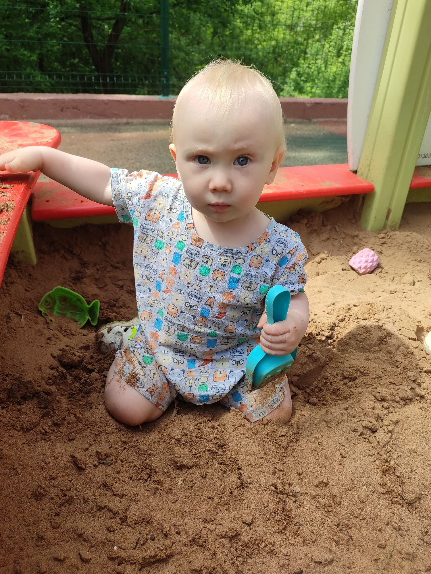 The image depicts a toddler, around one to two years old, playing in a sandbox. The child has fair skin, blond hair, and blue eyes. The toddler is wearing a patterned romper with short sleeves and shorts, featuring illustrations of animals and other objects. The child is kneeling in the sand, holding a blue plastic toy shovel in one hand and resting the other on the red edge of the sandbox. Several other plastic toys are visible in the sand, including a green fish mold and a pink textured ball. The sandbox is partially constructed with red, green, and white plastic elements. The background shows greenery, suggesting the sandbox is located in a park or playground. The sandbox appears to be in a city park in Izhevsk, Russia, based on the background and the style of playground equipment. The toddler's legs are partially buried in the sand, and there are patches of sand on the toddler's clothing. The expression on the toddler's face is serious and focused.