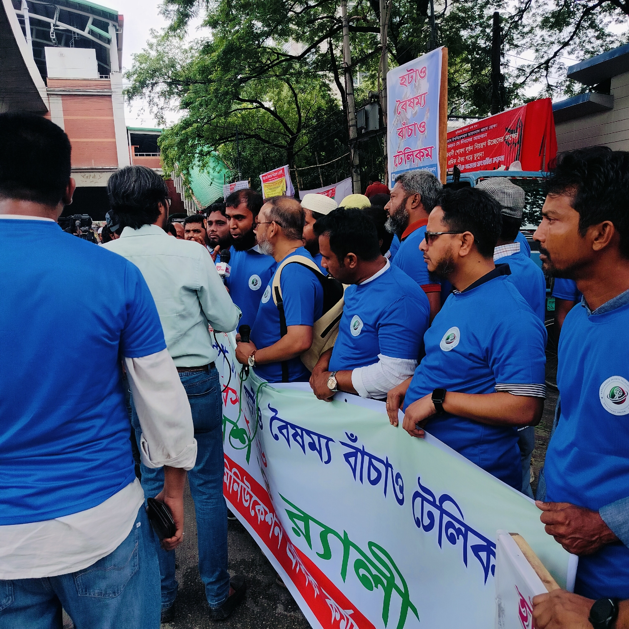 A medium, eye-level shot shows a rally or demonstration in an urban environment. A group of men, mostly facing forward, stand together, holding a large banner with text written in Bengali. They are uniformly dressed in blue t-shirts, some with white collars and cuffs, and each t-shirt has a small emblem on the left breast.
The banner they hold is light green, with dark blue and green writing. The text is largely unreadable due to the angle and distance.
To the left, a man in a light-colored shirt and blue jeans stands with his back to the camera. He appears to be interacting with the group of men holding the banner, possibly speaking to them. Behind him, a news crew or cameraman appears to be capturing the event.
In the background, signage on poles is visible, along with trees and a building constructed of red brick. A red banner is also seen above the heads of some of the people holding the banner. The location is the city of Unknown, Unknown.