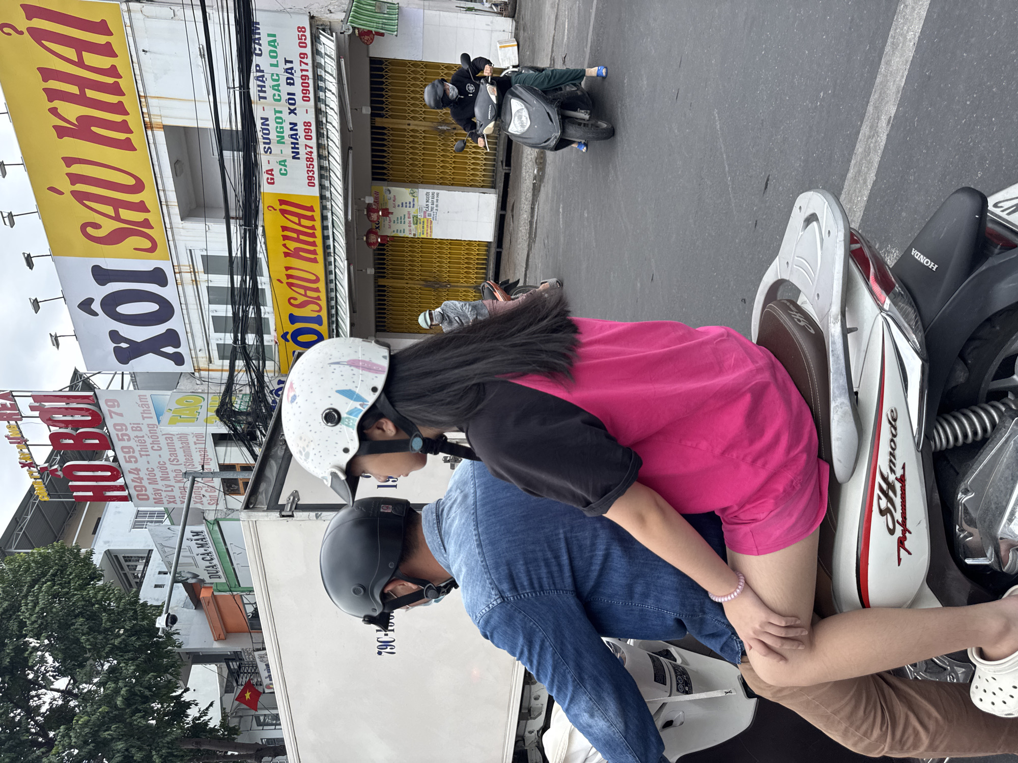 The image depicts a street scene in Nha Trang, Vietnam. In the foreground, a person is riding on the back of a white scooter, wearing a pink shirt and shorts, and a white helmet with a decorative pattern. They are holding onto the driver, who is wearing a blue denim shirt and a black helmet. The scooter has "SH mode" branding on its side.
In the background, there are several buildings with signs in Vietnamese. One prominent sign, in large red and yellow lettering, reads "XÔI SÂU KHAI." Another sign is partially visible with the text "HO BOI." Various other signs and advertisements are present on the buildings, along with electrical wires and some greenery.
To the right of the main scooter, another person is riding a dark-colored scooter, wearing a dark jacket and pants, a black helmet, and a face mask. They are also wearing blue sandals. Further in the background, another person is walking on the sidewalk, wearing a light-colored hoodie and a light-colored helmet. The street appears to be paved, and there are some traffic markings visible. The overall impression is of a busy urban environment in Vietnam.