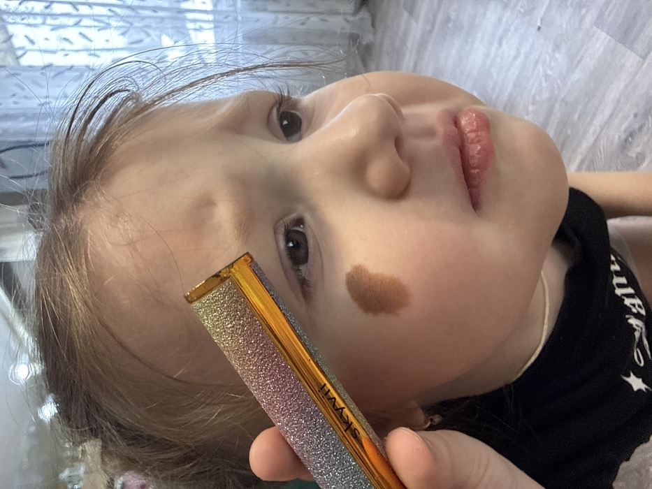 An indoor close-up photograph taken in the city of Кипчак, Russia, depicts a young, light-skinned child with a furrowed brow and a brown, irregular mark on their left cheek. The child is wearing a black garment and a light-colored necklace. An adult hand, visible in the lower left, holds a cylindrical cosmetic item marked "skyvii," featuring a glittery, multicolored casing with an amber tip, positioned diagonally near the child's left eyebrow. The child's gaze is directed downwards, and their mouth is slightly agape. The background consists of light gray laminate flooring and a window covered by a sheer white curtain.