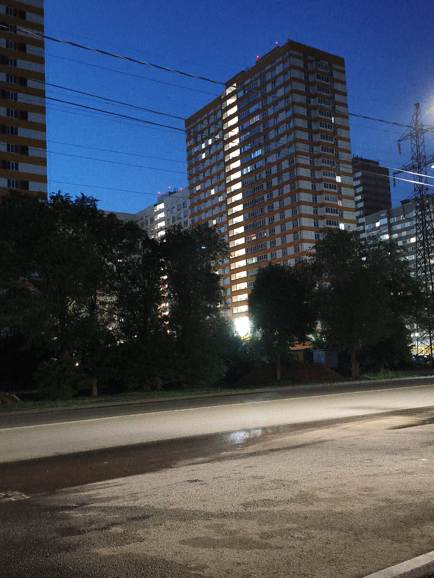 The image depicts a nighttime street scene in Orenburg, Russia. In the background, several high-rise apartment buildings are visible, with many windows illuminated, suggesting the time is evening or night. The sky is a deep blue, indicating it is twilight or after dark. A large tree-lined area is in the mid-ground, obscuring parts of the buildings. In the foreground, a wet asphalt road is visible, reflecting some light. Overhead power lines stretch across the frame. A tall metal electrical pylon is also present on the right side of the image. The overall impression is of an urban environment at night, likely in a residential area.