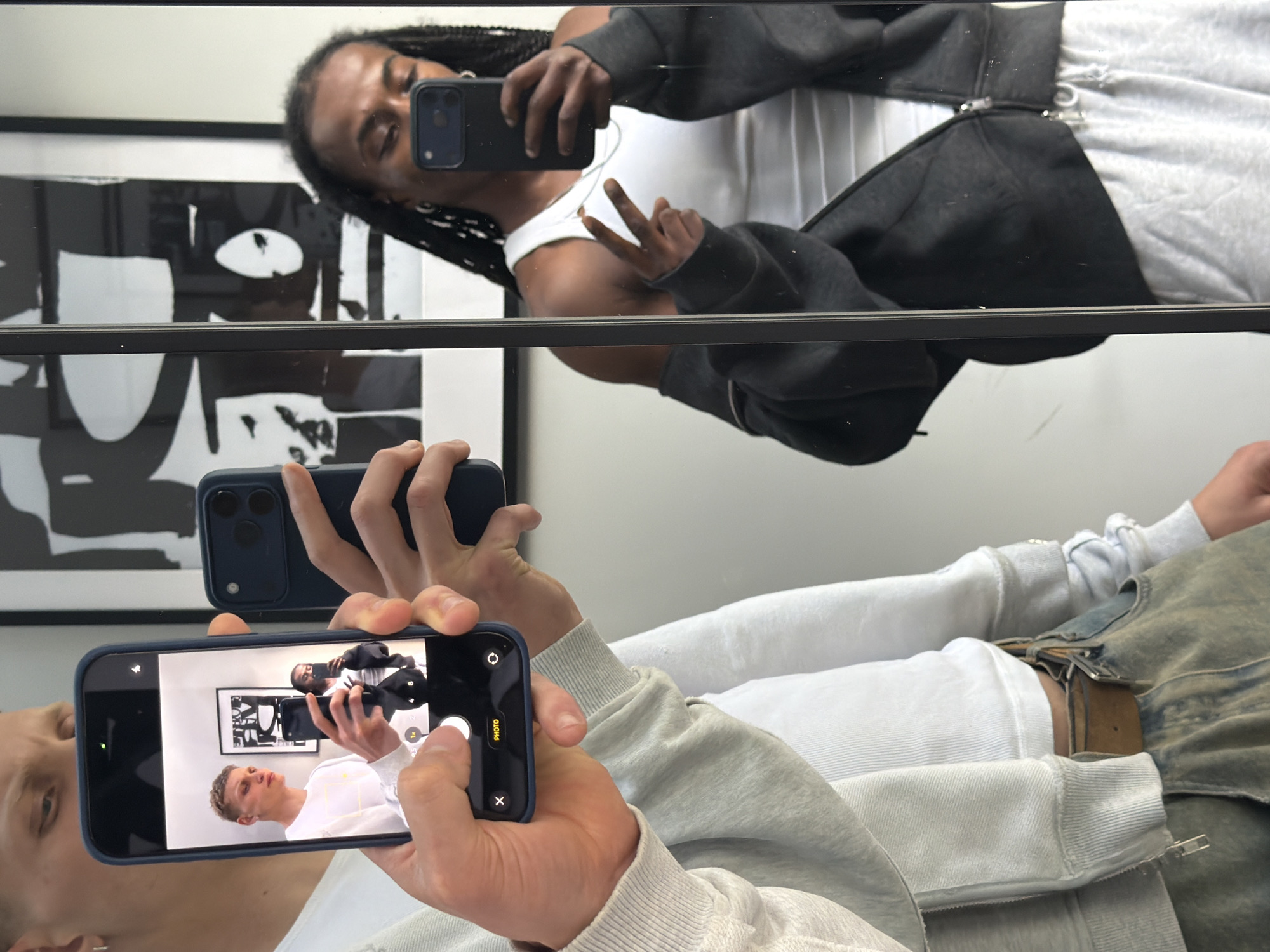 Two individuals are posing for a selfie reflected in a mirror, which is positioned in a well-lit interior space. The person on the left, holding a dark blue smartphone, is capturing a photo of themselves and another individual. This second individual is visible in the phone's screen, also holding a phone and taking a selfie. Both individuals are wearing casual attire. The person on the right, whose reflection is clearly visible in the mirror, is wearing a white tank top and a dark grey zip-up hoodie, with their hair styled in braids. They are making a peace sign with their fingers. The background includes a framed piece of abstract art on the wall behind them. The environment appears to be a modern indoor setting, possibly a home or a studio, given the clean walls and the presence of artwork. There is no information in the image to suggest a specific location within the city of London, United Kingdom.