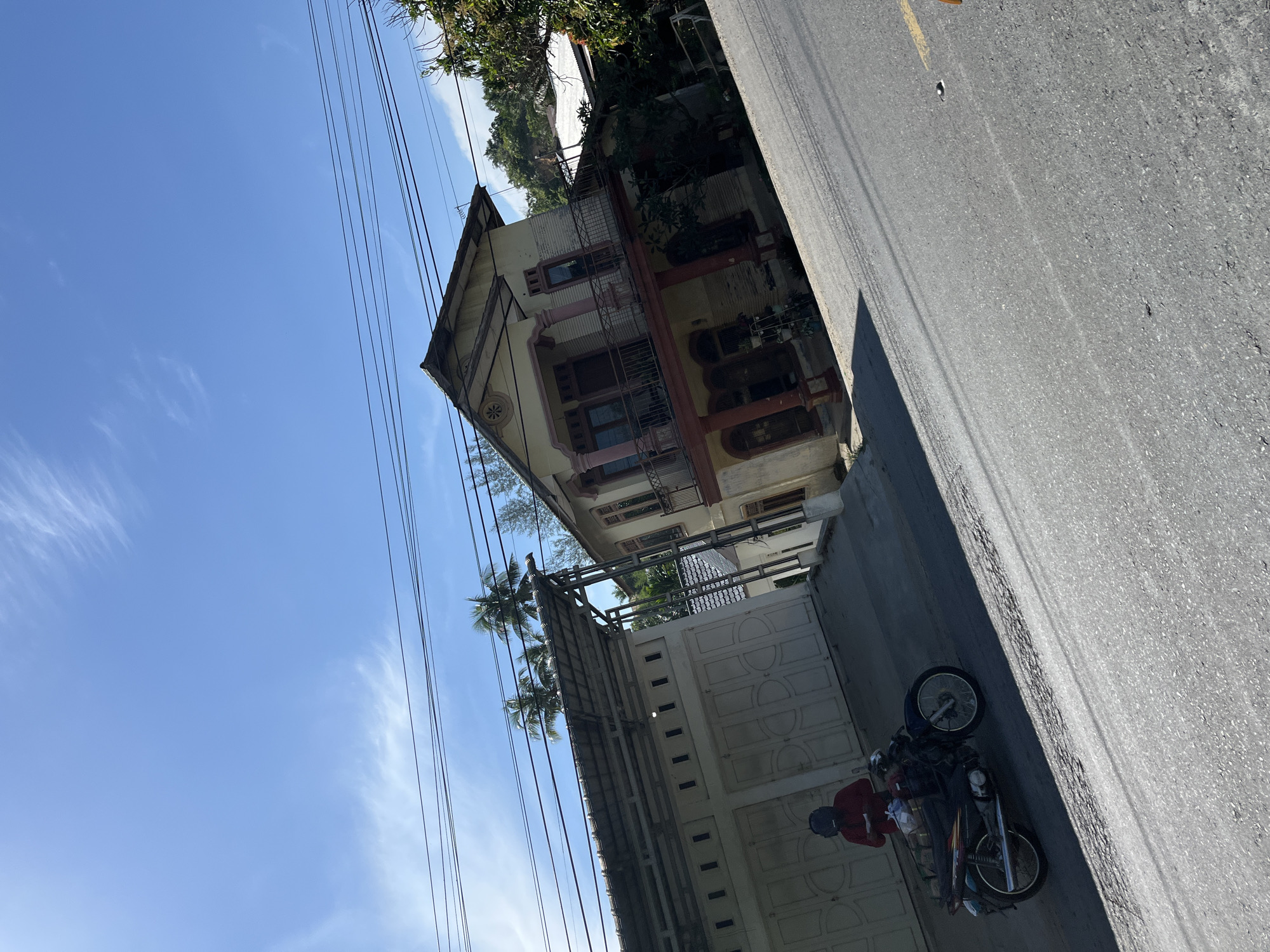 The image depicts a street scene in a residential area, likely in Bukitrata, Indonesia. A weathered, multi-story building with a pale yellow facade and reddish-brown trim stands prominently on the left. The building features balconies with decorative railings and arched windows. Adjacent to the building is a white gate with a patterned design, and a covered walkway or carport. Tropical trees, including palm trees, are visible, suggesting a warm climate. Overhead, a network of power lines stretches across the bright blue sky, which has wisps of white clouds. The road in the foreground is paved and appears to be asphalt.
A motorcycle is parked on the right side of the road, with its kickstand down. A person wearing a red shirt and a helmet is seated on the motorcycle, facing away from the camera. The scene appears to be set during the daytime, with bright sunlight casting shadows, indicating it could be late morning or early afternoon. The overall atmosphere is casual and everyday, portraying a typical street view in a tropical locale. No visible text is present in the image.