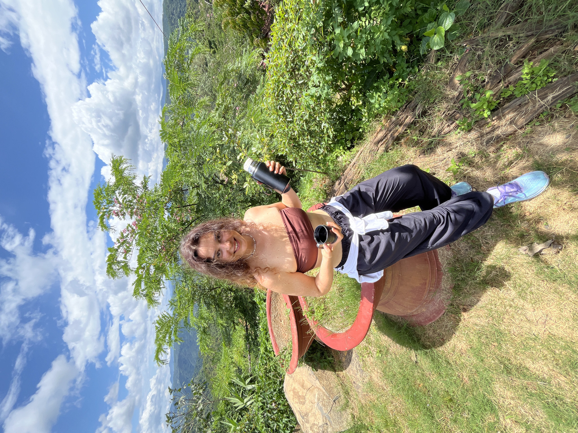 A young Caucasian woman with curly brown hair and a tan complexion is pictured outdoors on a sunny day, holding a black thermos in her left hand and a small black cup in her right. She is wearing a brown bandeau top, black sweatpants tied with a white shirt around her waist, and light blue athletic shoes with purple laces. She is seated on the edge of a large, reddish-brown planter with a textured surface, which is positioned on a grassy area. The planter appears to be made of clay or concrete and contains sparse green foliage. In the background, lush green vegetation covers rolling hills under a bright blue sky with white, fluffy clouds. The scene is set in Sơn Điền, Vietnam, characterized by its tropical landscape. The woman is smiling directly at the camera.